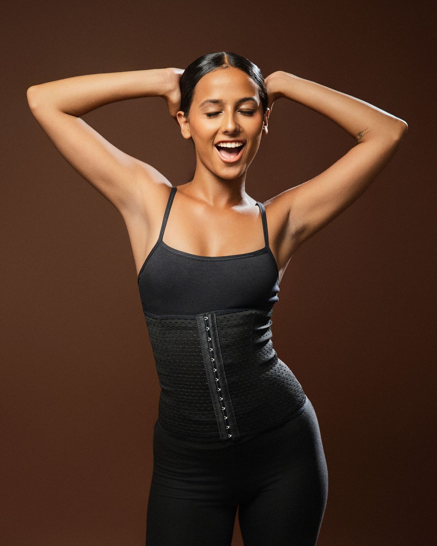 Athletic wear model demonstrates black waist trainer against brown studio background.