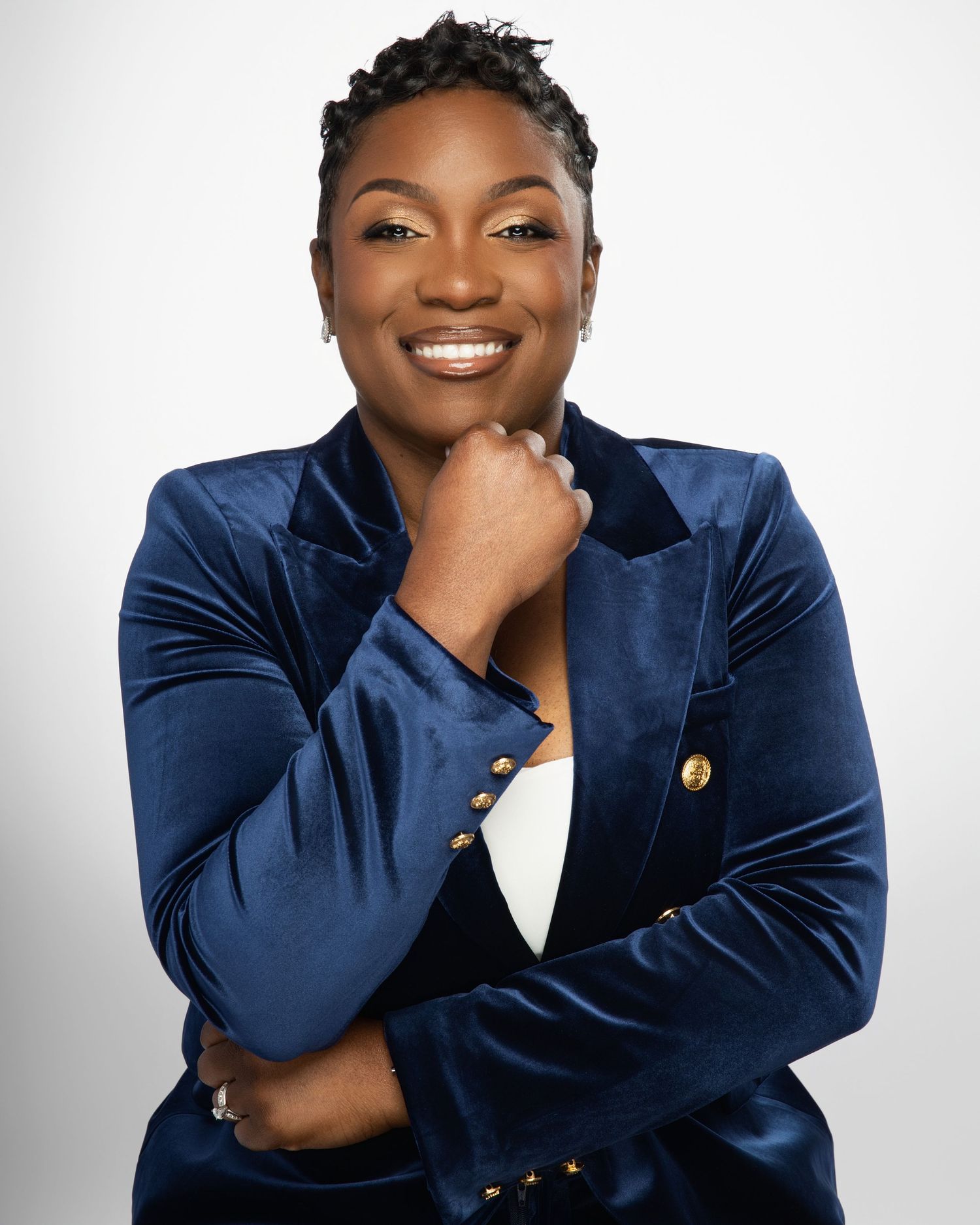 Professional in navy velvet blazer poses with hand on chin against white background.