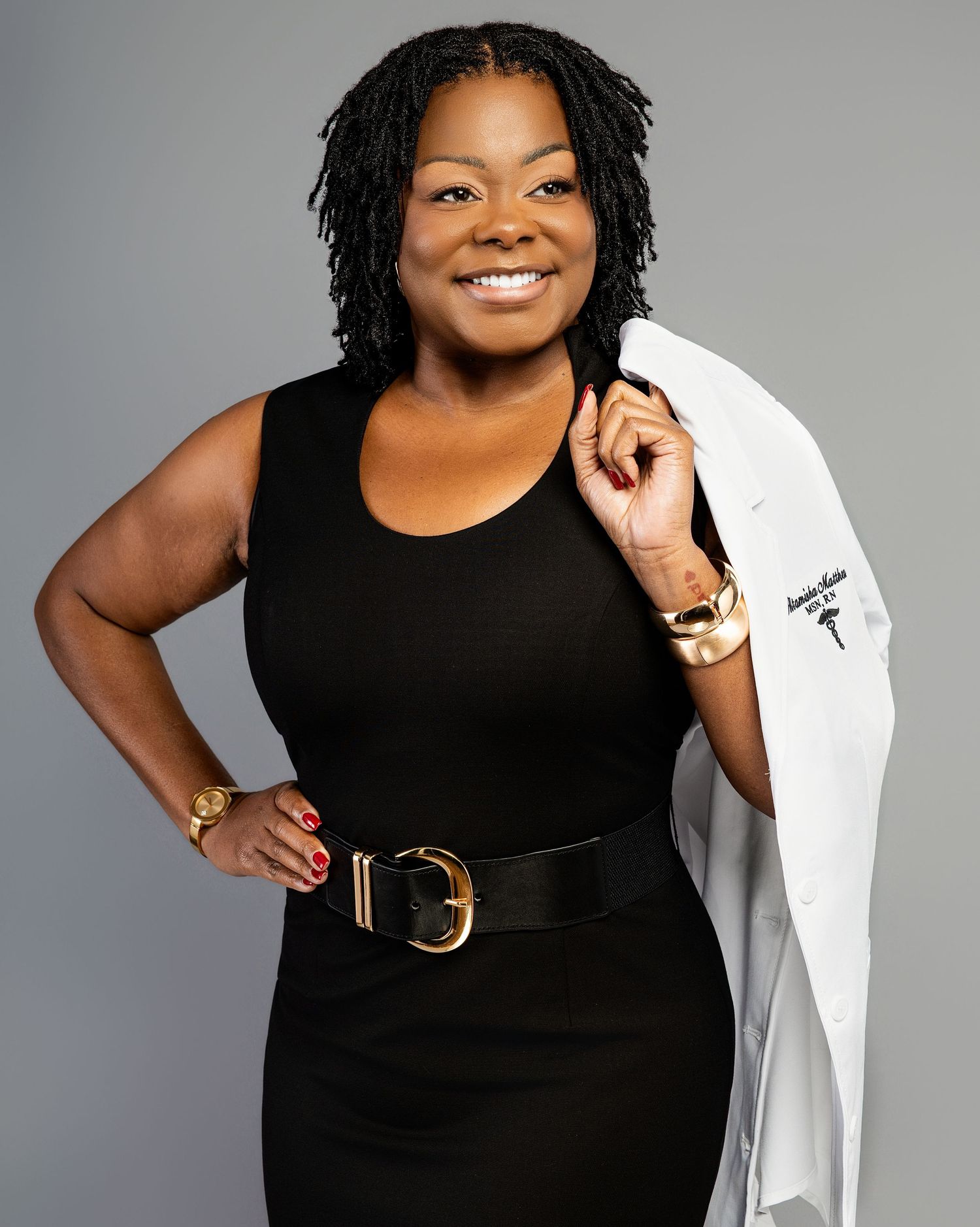 Medical professional in a black sleeveless dress and white coat poses confidently against gray background.