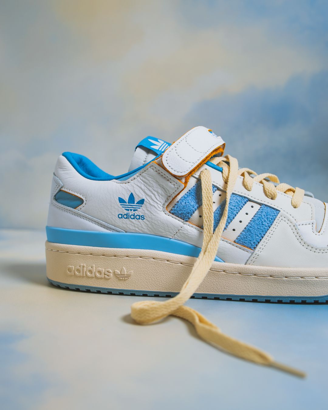 Classic Adidas Forum sneaker in white and light blue colorway with cream laces photographed against cloudy background.