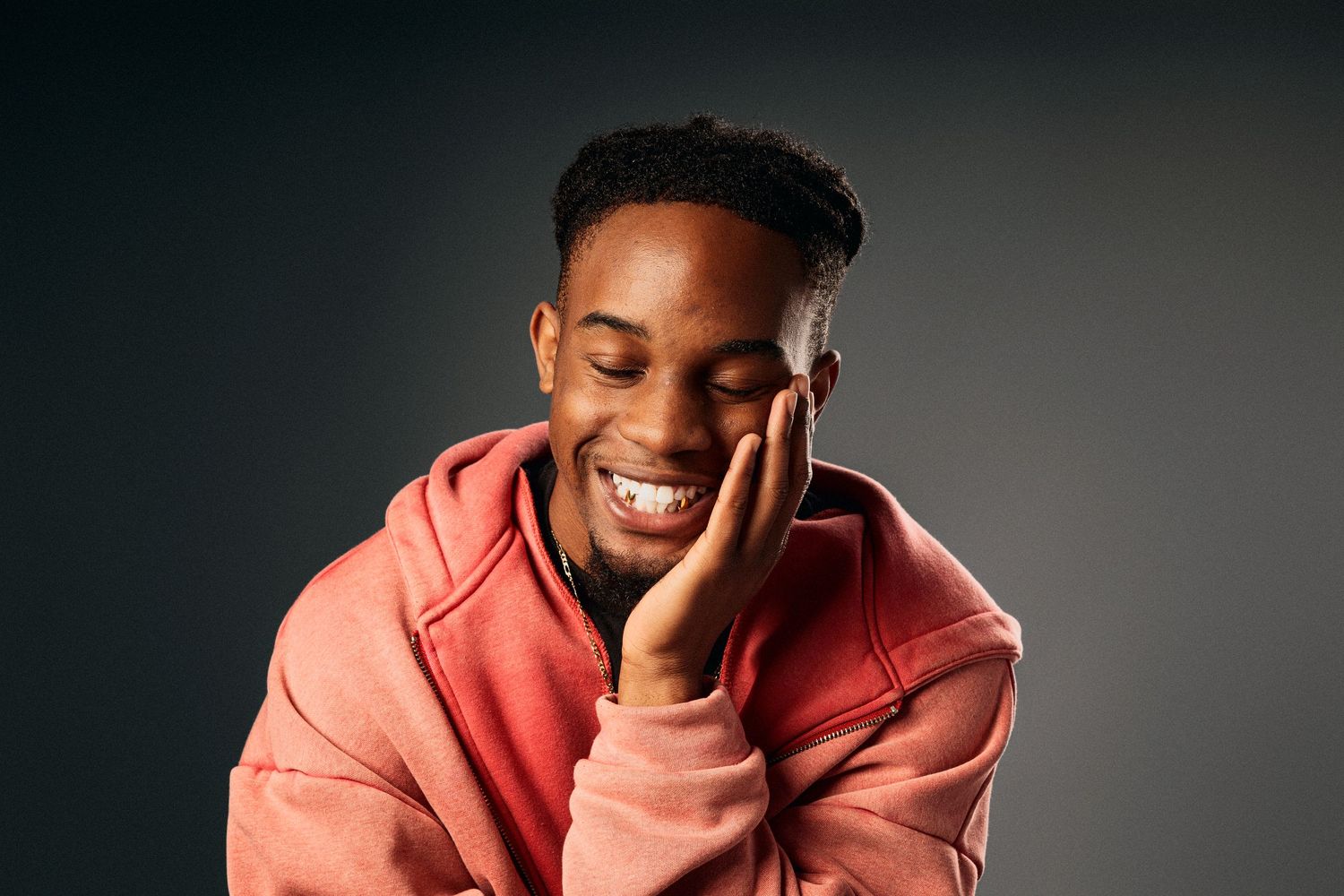 A person wearing a coral colored hoodie smiles while posing against a dark background in a photo series.