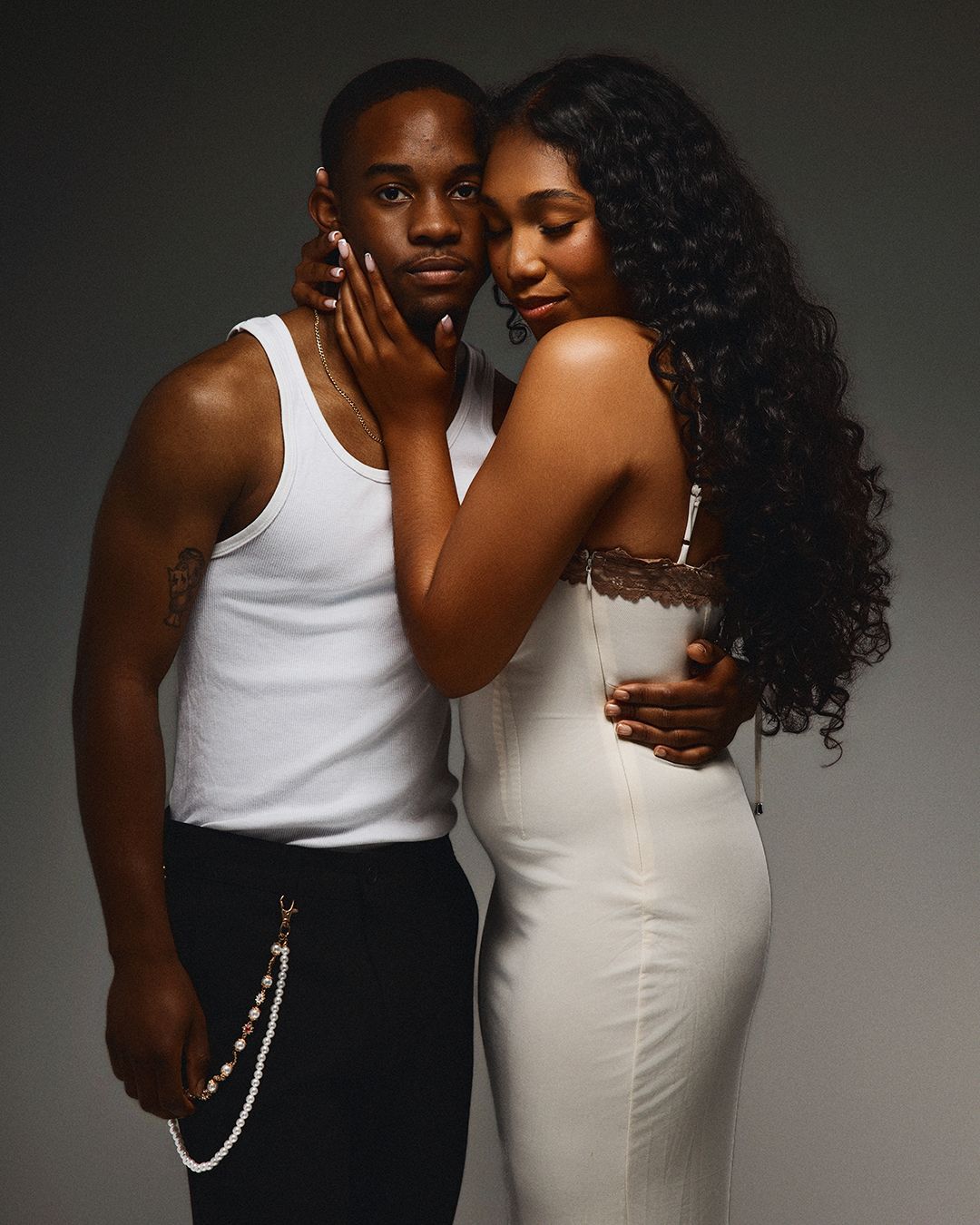 A young Black couple poses intimately in matching white outfits against a gray studio background.