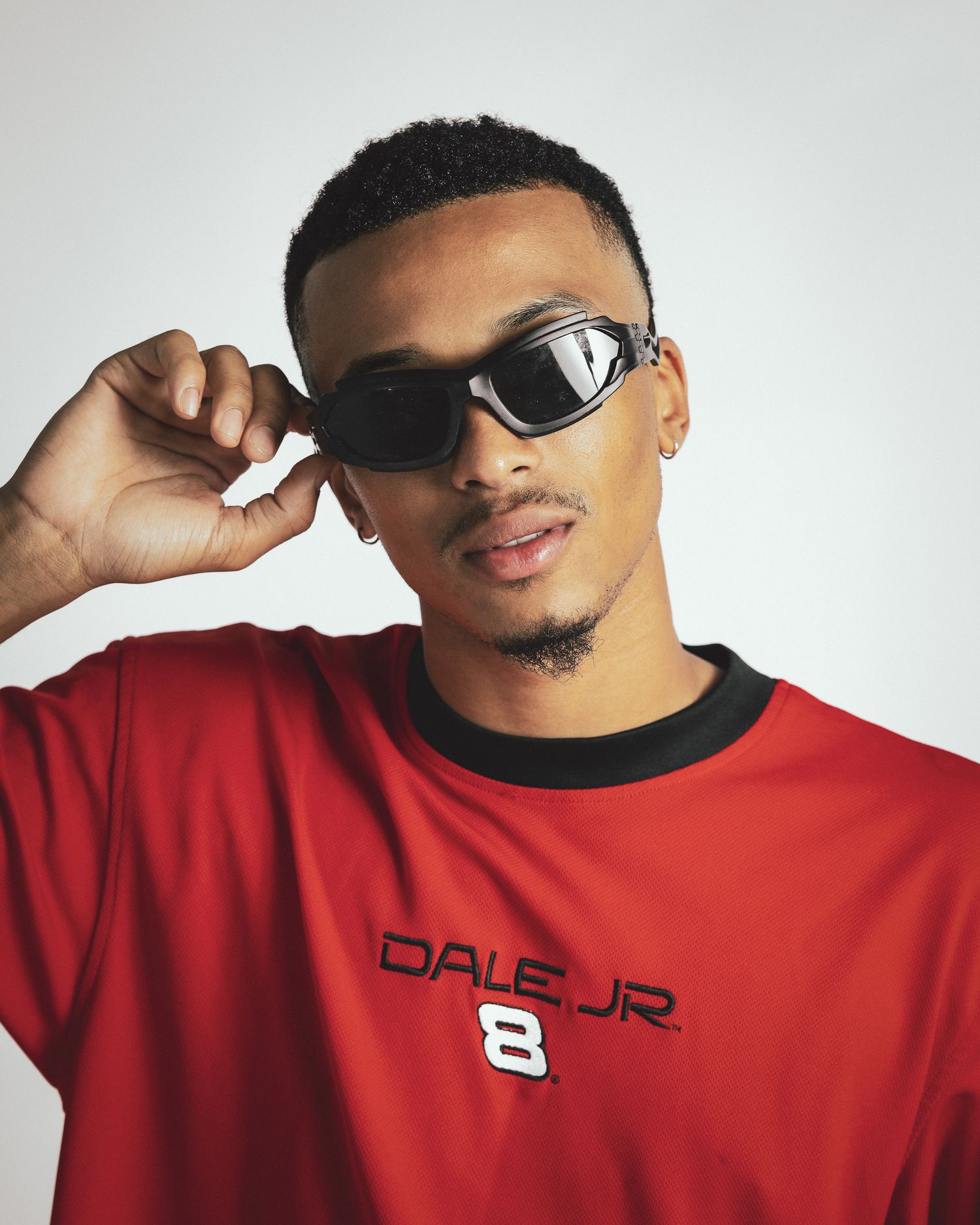 Person wearing sunglasses and a red Dale Jr. 8 shirt poses against white background while adjusting eyewear.