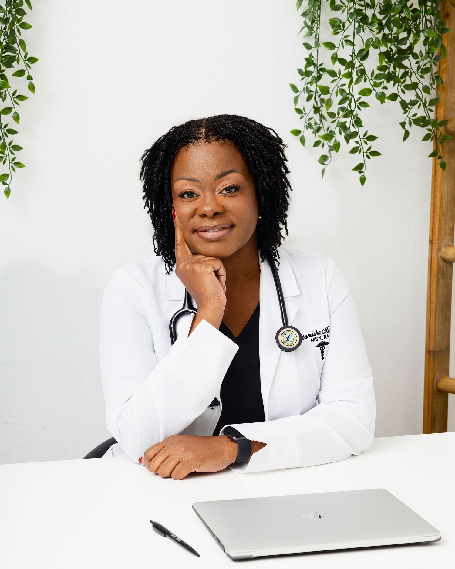 Healthcare professional in white coat sits at desk with laptop and stethoscope near decorative hanging plants.