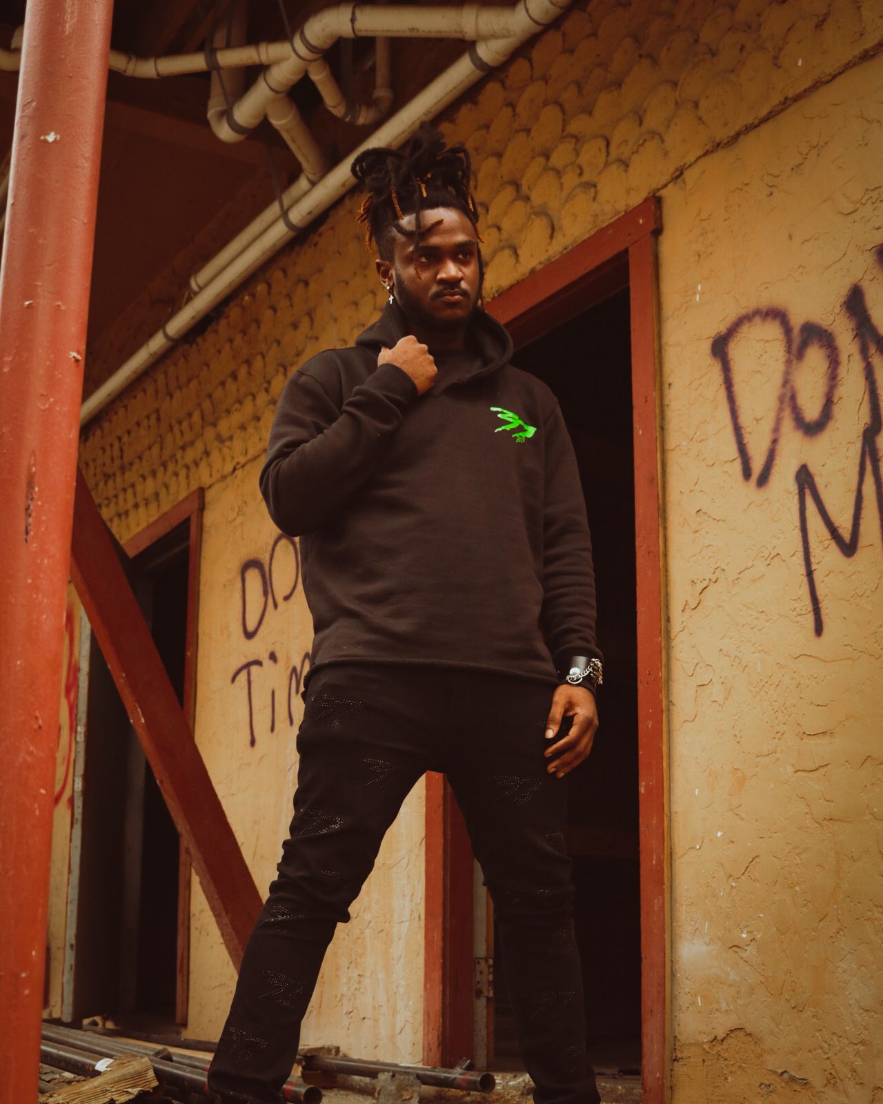 Someone in a black hoodie with a small green logo poses against a graffiti-covered wall in an urban setting.
