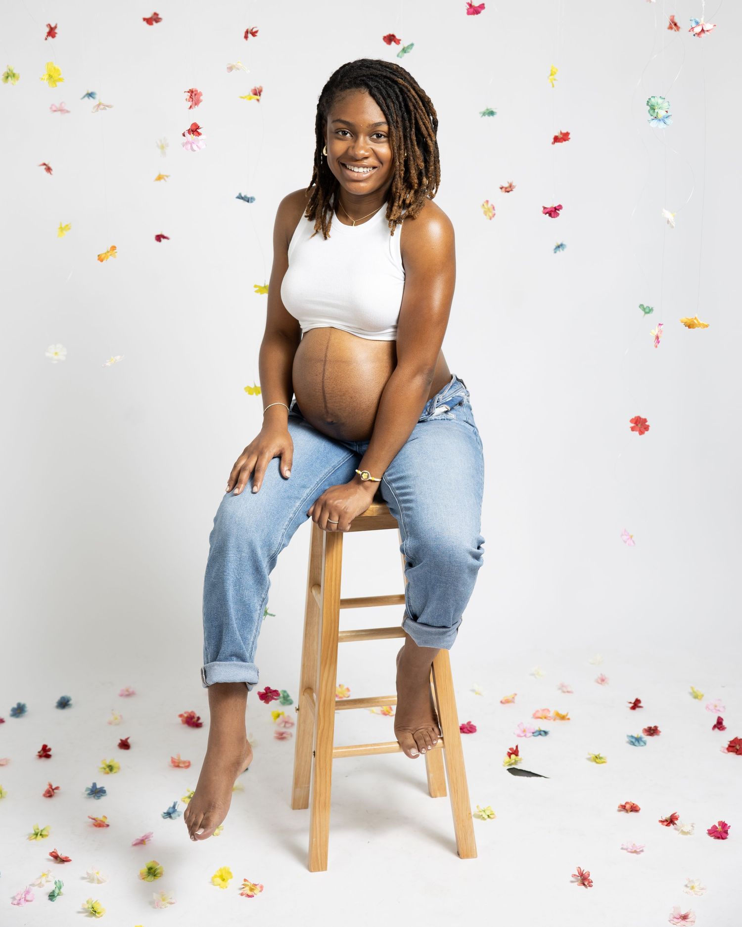 Expectant person in white crop top and jeans sits casually on wooden stool with colorful confetti background.