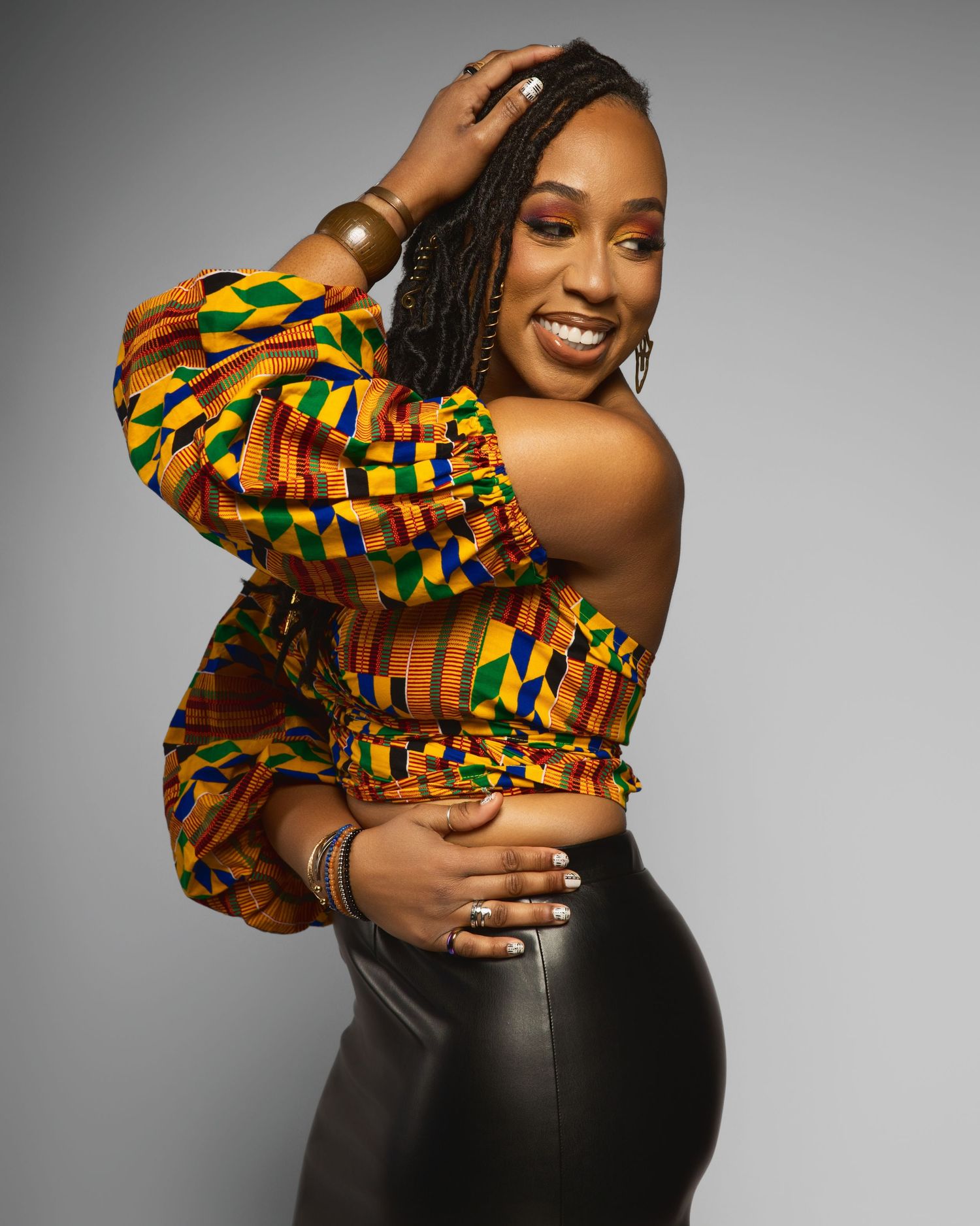 Model poses in vibrant African print crop top with puff sleeves and black leather skirt against grey background.