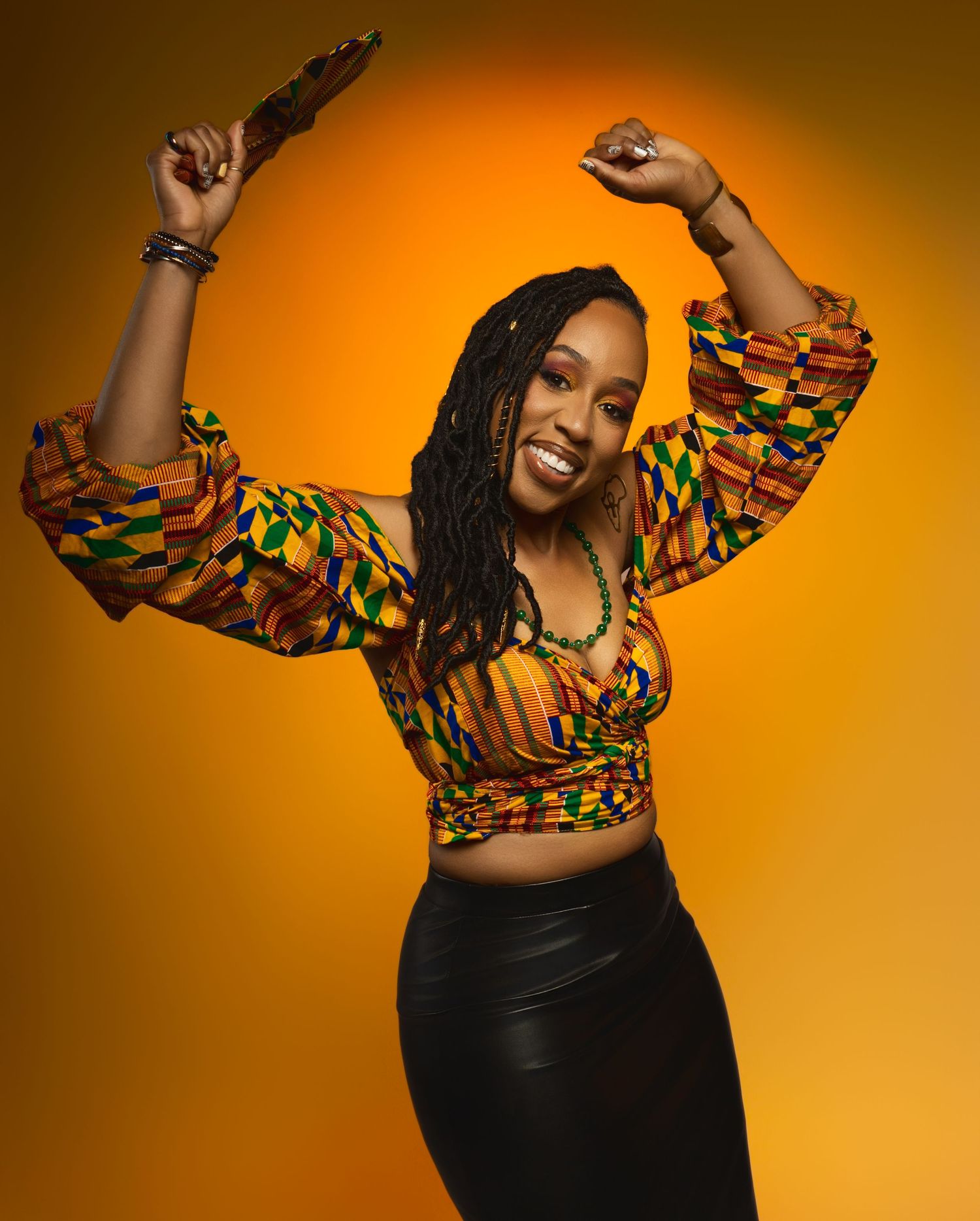 Dancing pose in African print crop top and black skirt against orange backdrop.