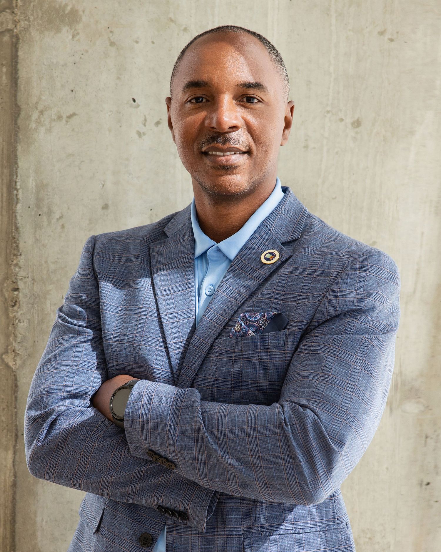 Professional wearing a gray plaid suit jacket poses against a concrete wall with arms crossed.
