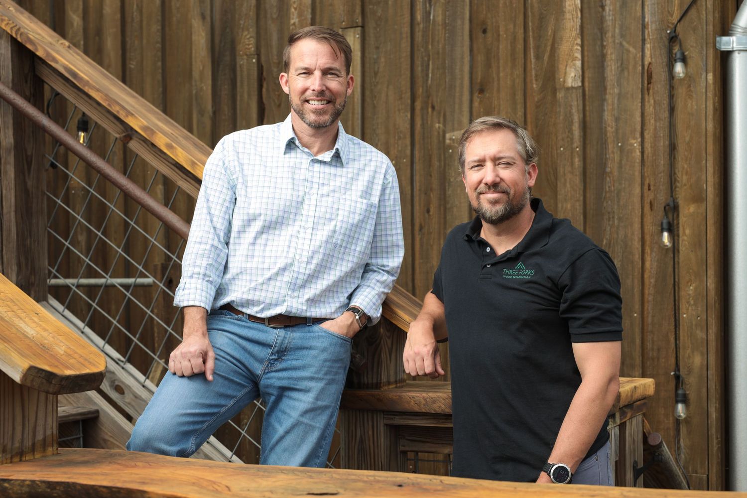 Two people in casual attire pose together on wooden stairs against a rustic wooden wall background.