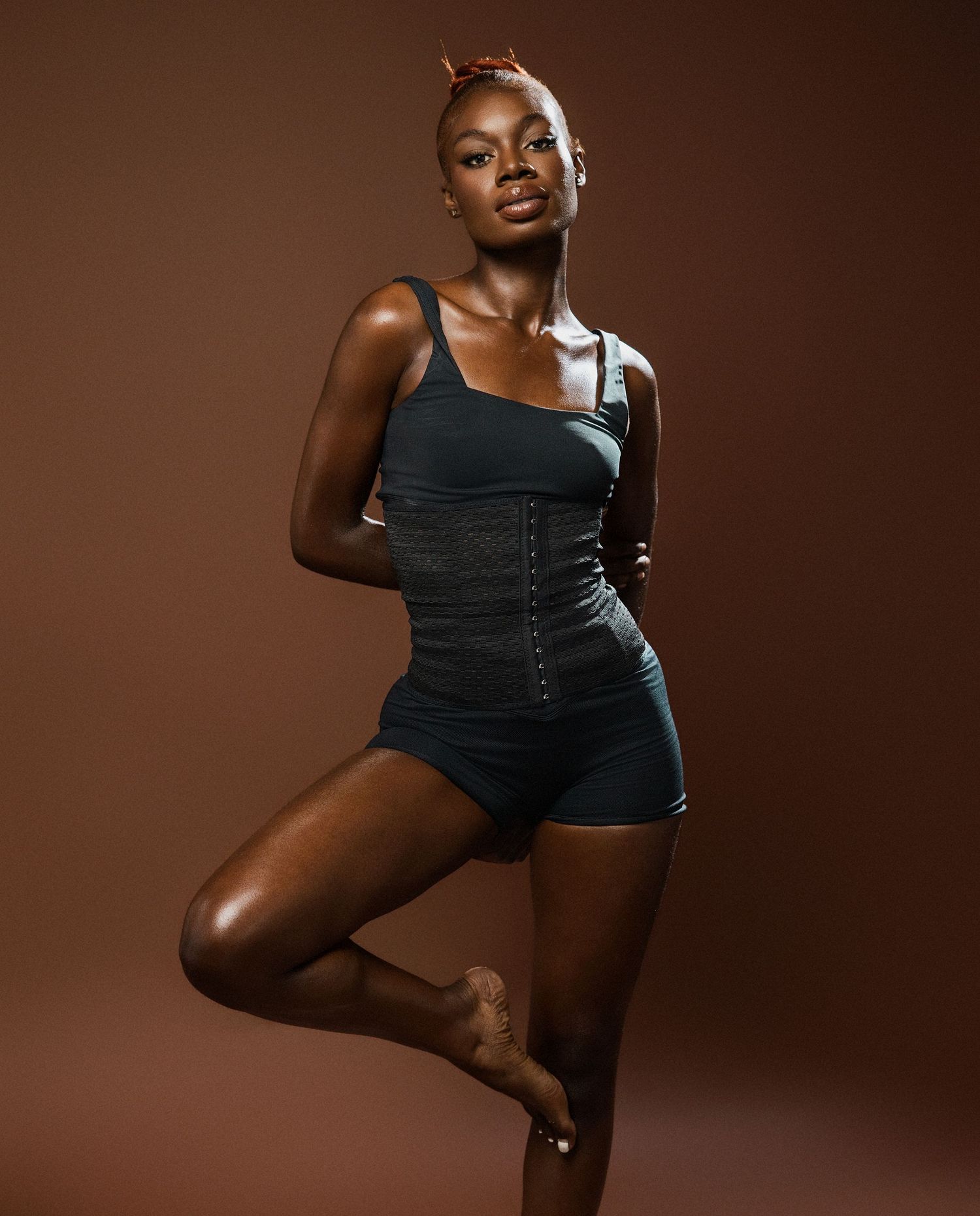 Athletic model in black workout gear poses dynamically against brown studio backdrop.