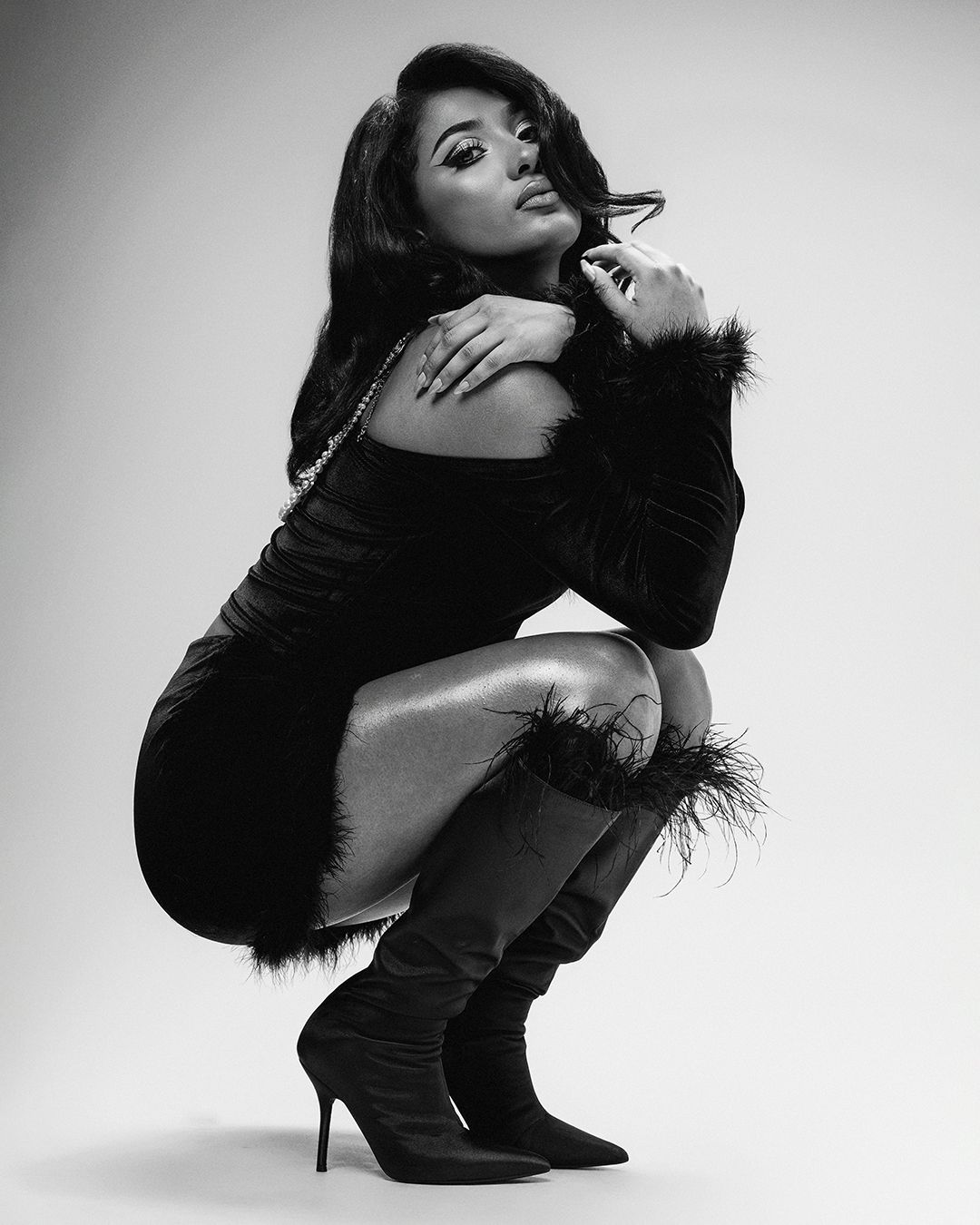 Dramatic black and white fashion photo of a model in a feather-trimmed black dress and boots posing in a crouched position.