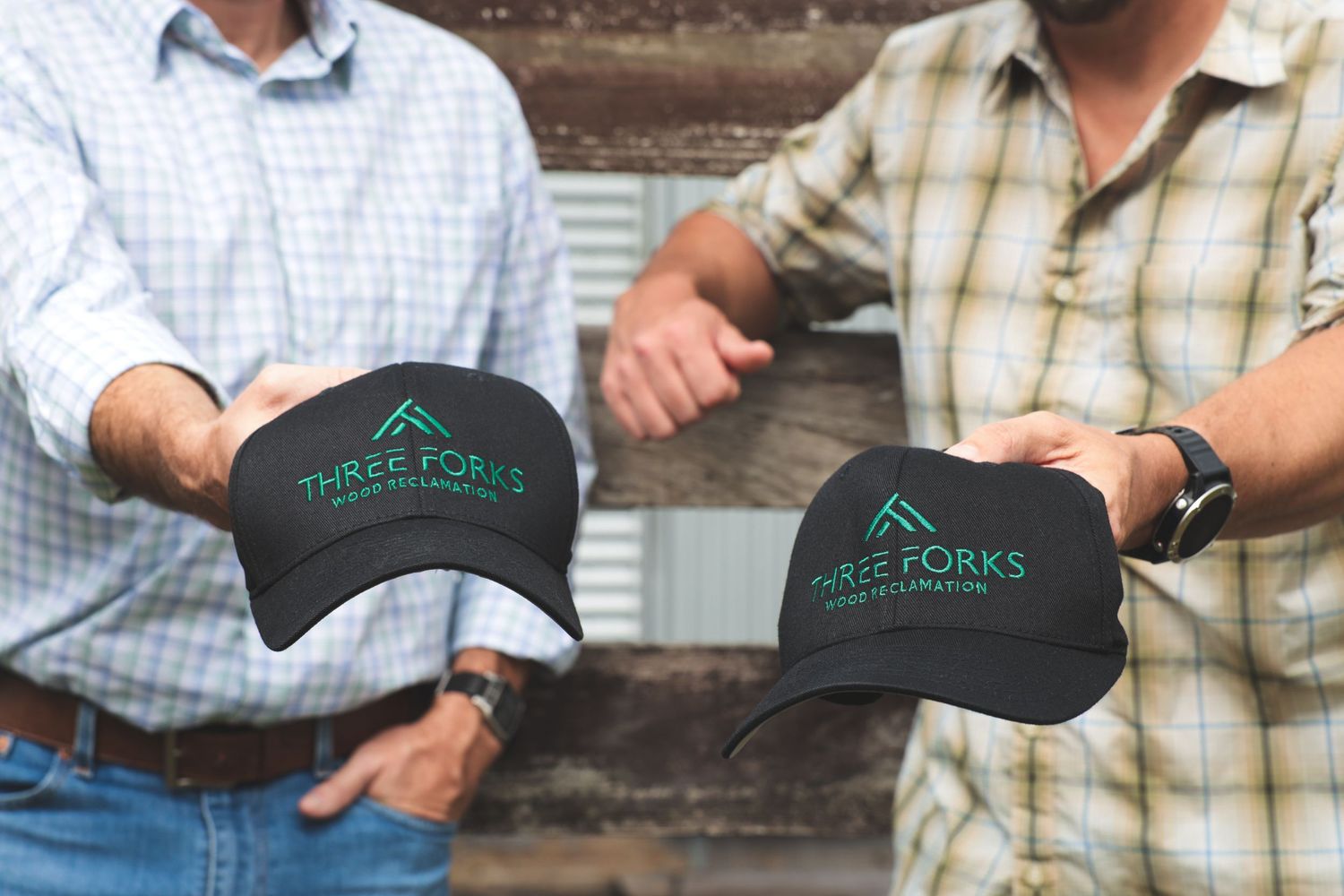 Two people wearing plaid shirts hold black baseball caps with teal logo embroidery against wooden background.