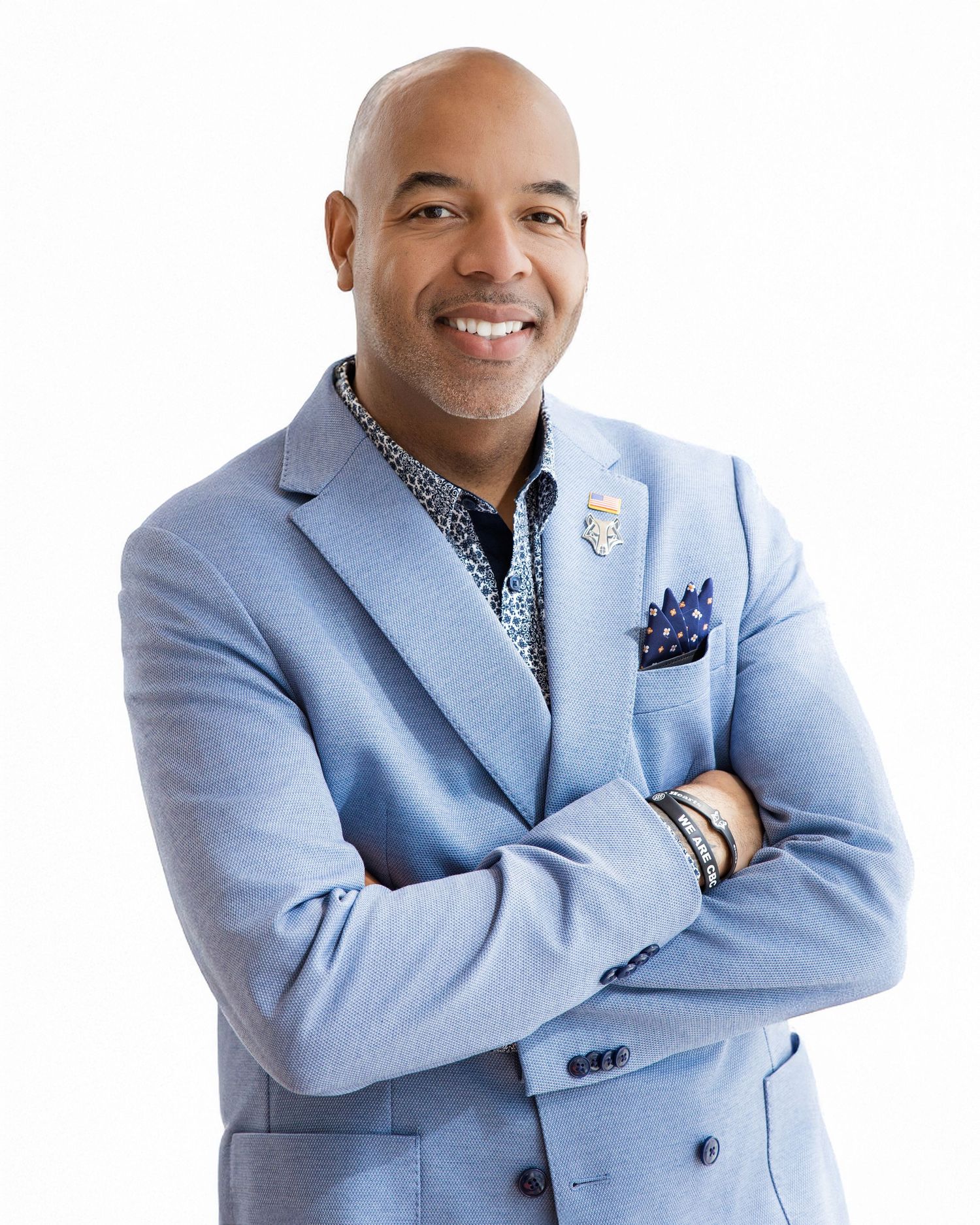 Professional wearing a light blue blazer and patterned shirt poses with arms crossed against white background.
