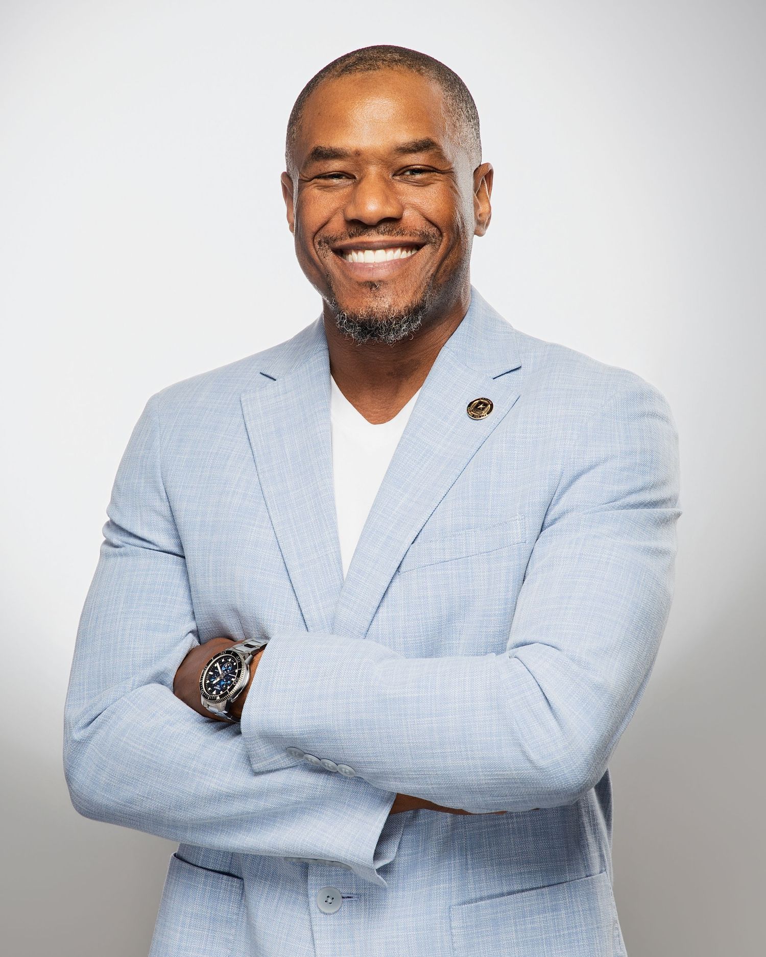 Smiling man in light blue blazer and white shirt, arms crossed, wearing a watch, professional headshot photo.