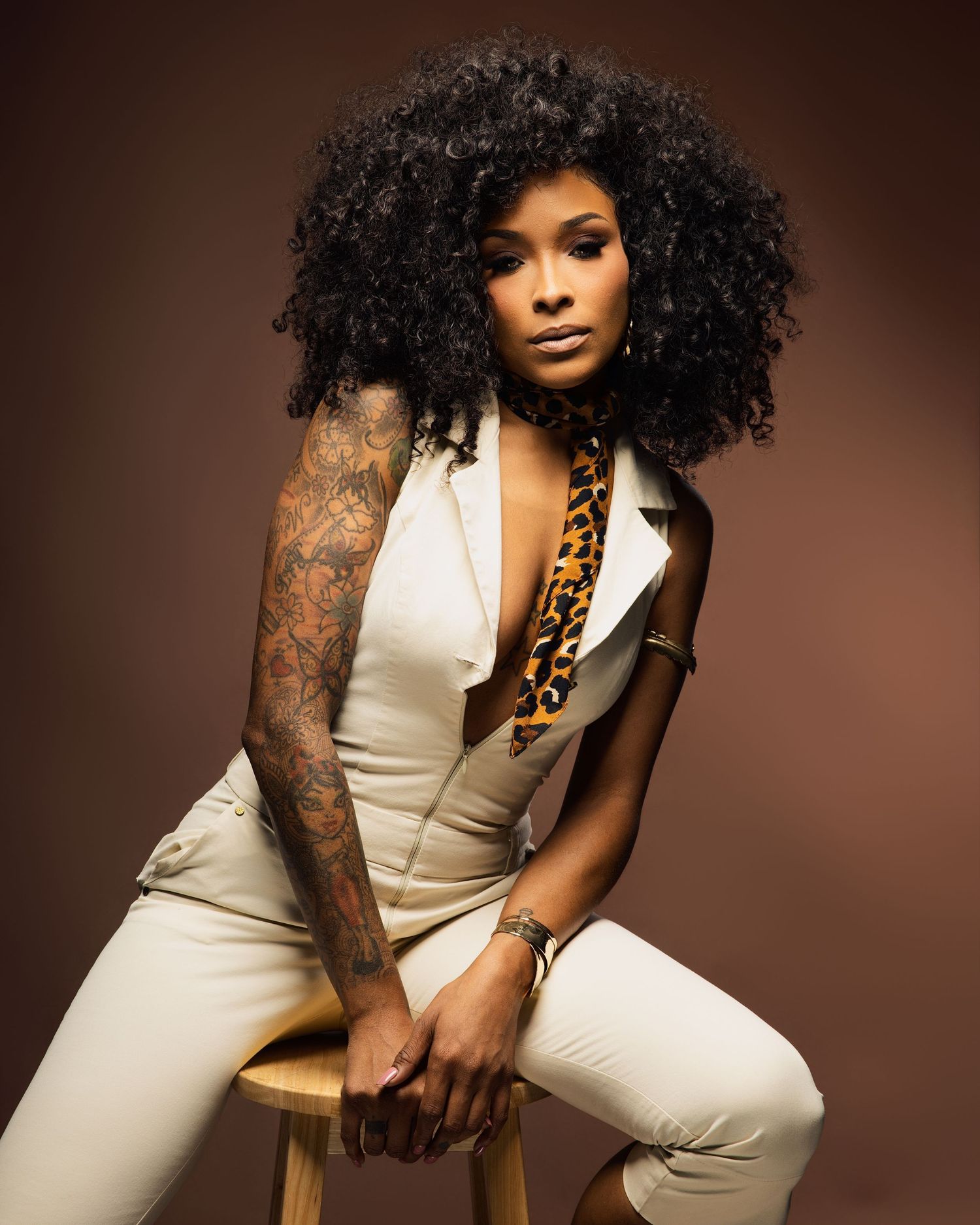 Model in white outfit with animal print details poses seated against brown backdrop.