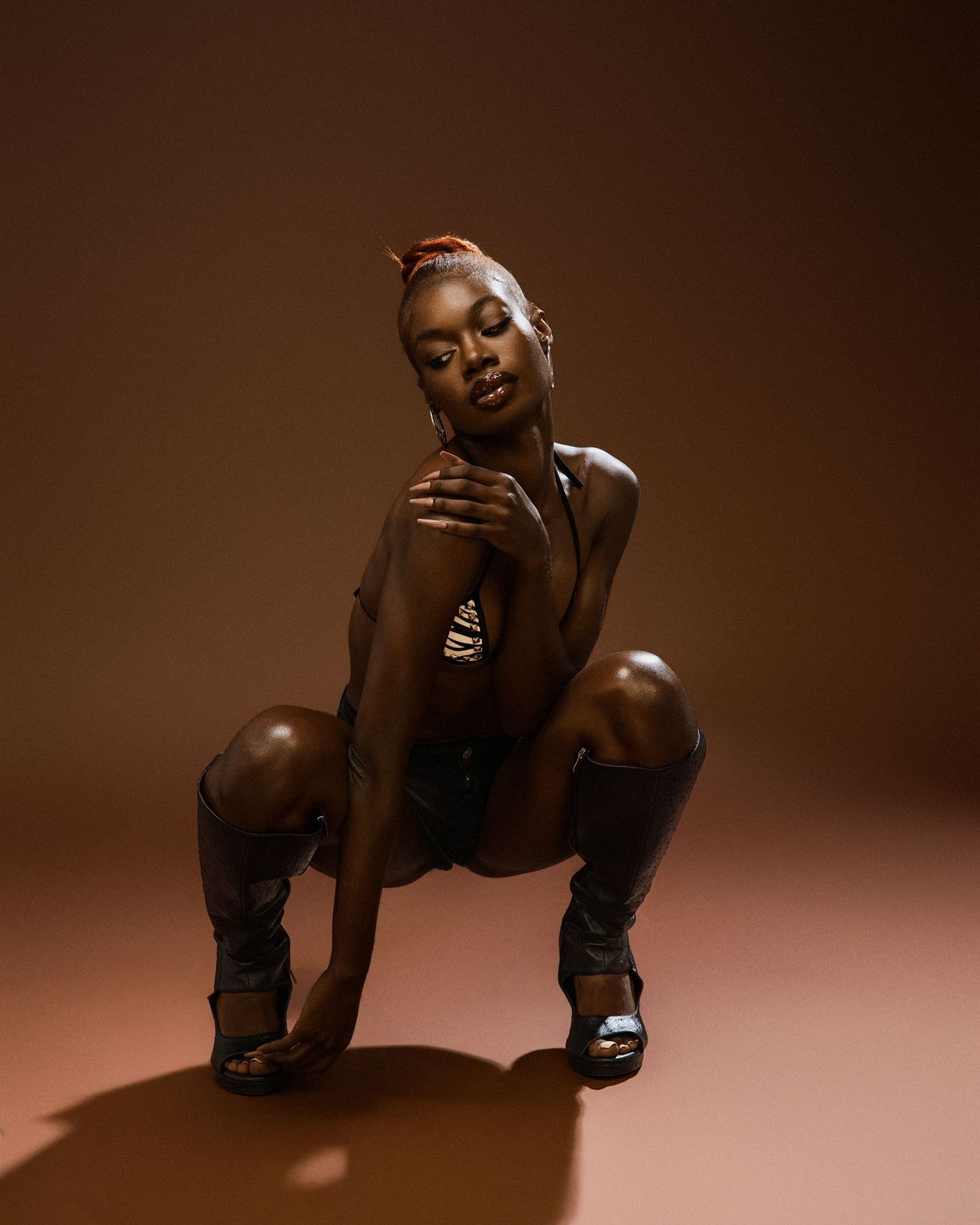 Artistic portrait of performer in black outfit crouching against brown studio background.