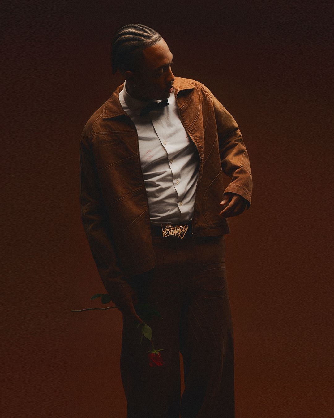 Figure in brown jacket and white shirt poses dramatically in moody lighting.