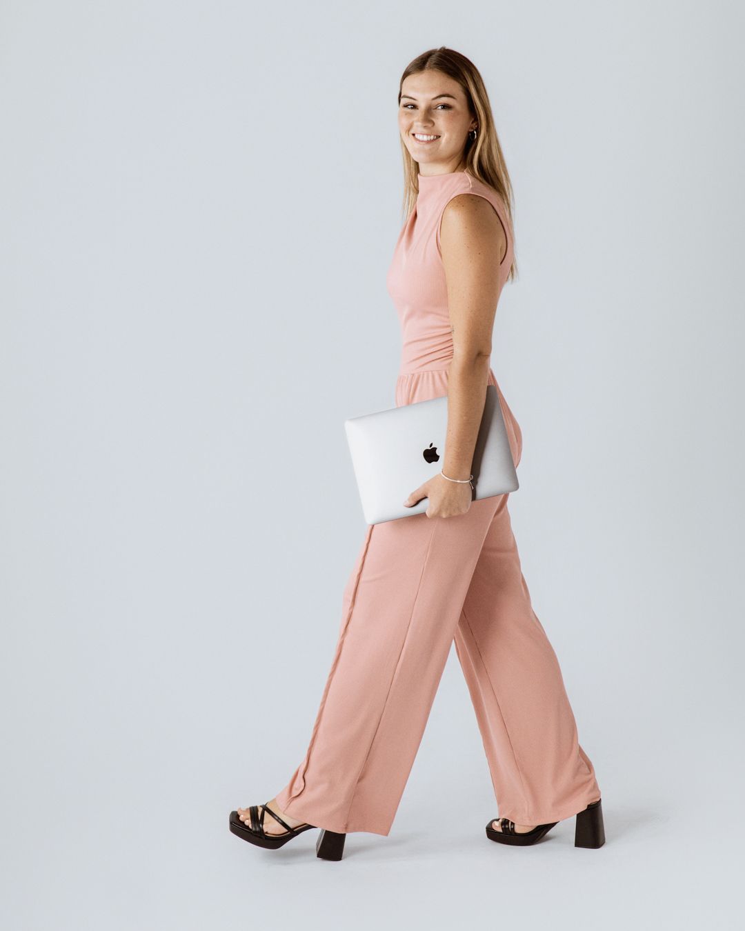 Woman in peach-colored jumpsuit poses with laptop in studio against white background.