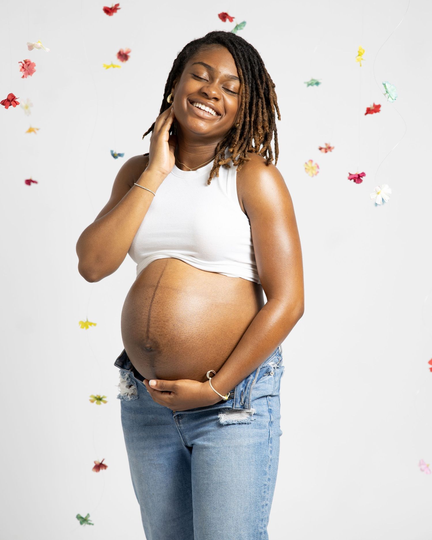 Pregnant woman in white crop top and jeans poses joyfully against white background with colorful confetti falling.