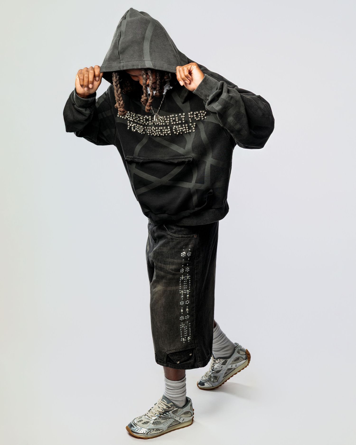 Person wearing a black oversized hoodie and shorts with grey sneakers shown from different angles in a studio setting.