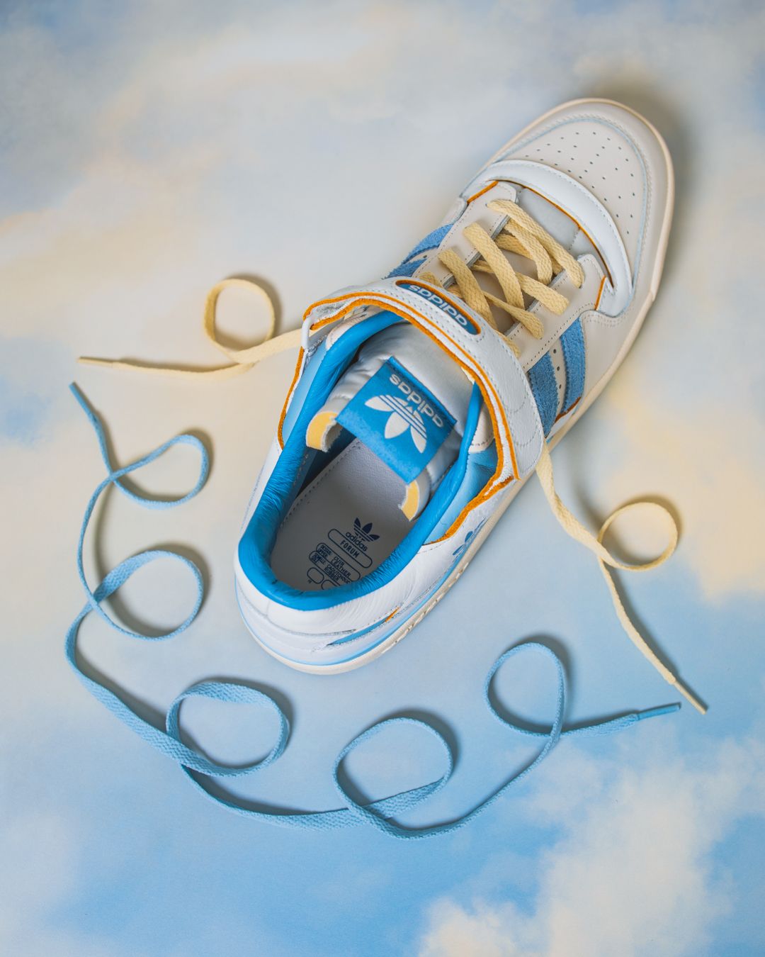 A white and blue athletic sneaker with untied laces photographed against a soft cloudy background.