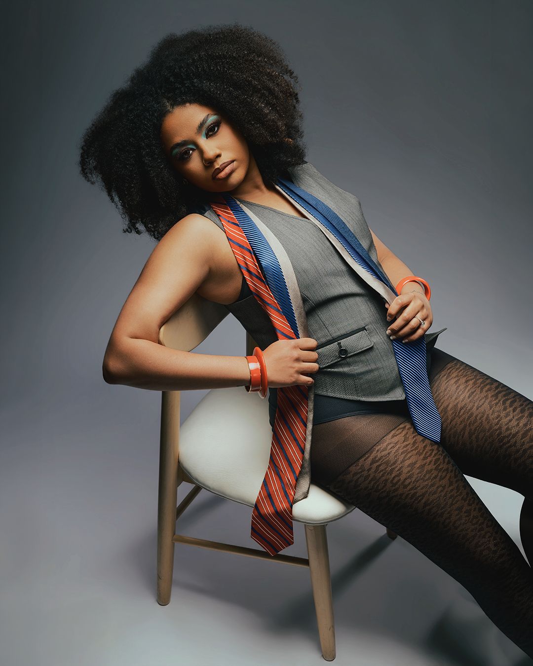 Woman with natural afro hair poses on stool wearing gray vest, blue blazer, and patterned tights with red tie.