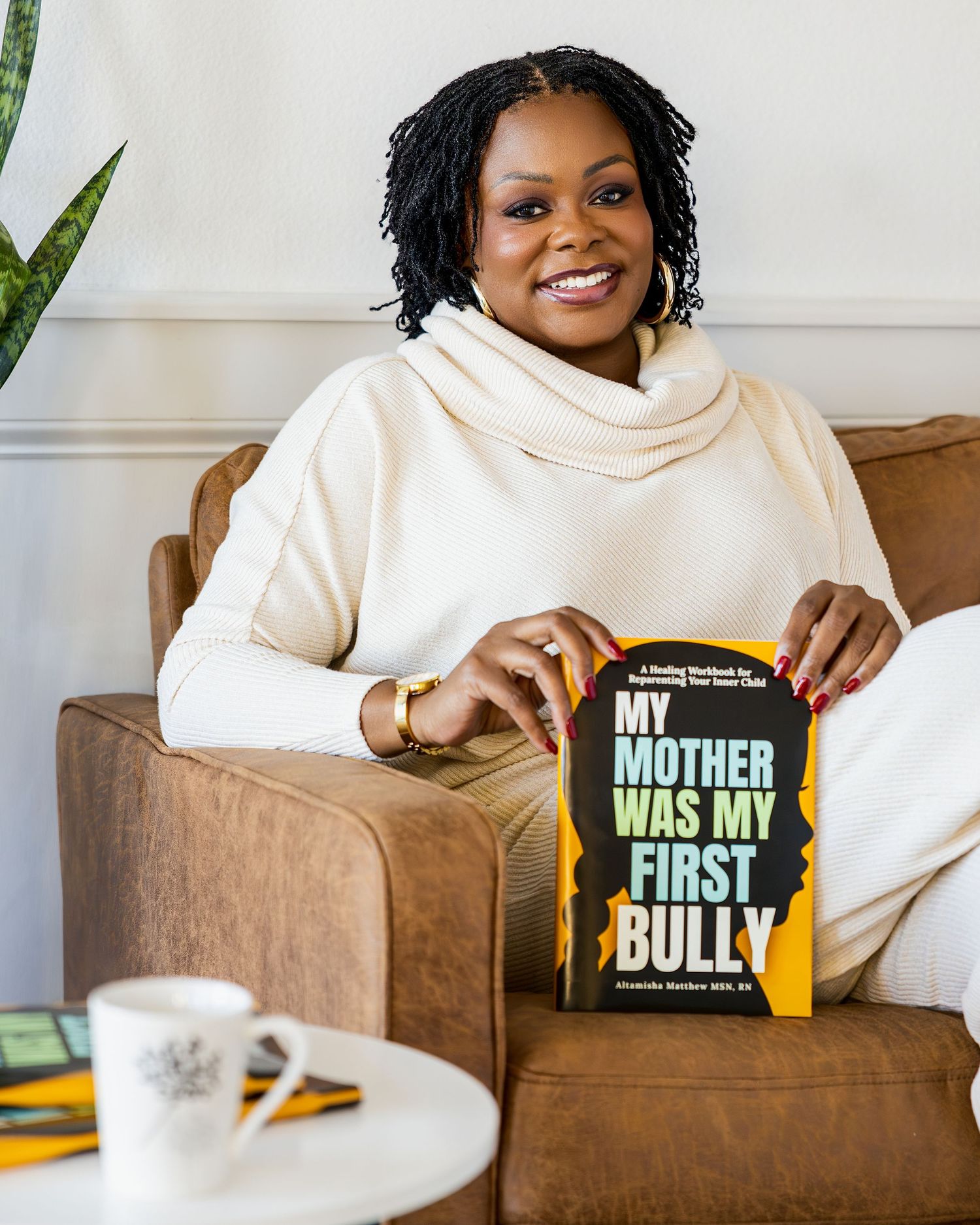 A person in a cream turtleneck sweater sits in a brown armchair holding the book 'My Mother Was My First Bully'.