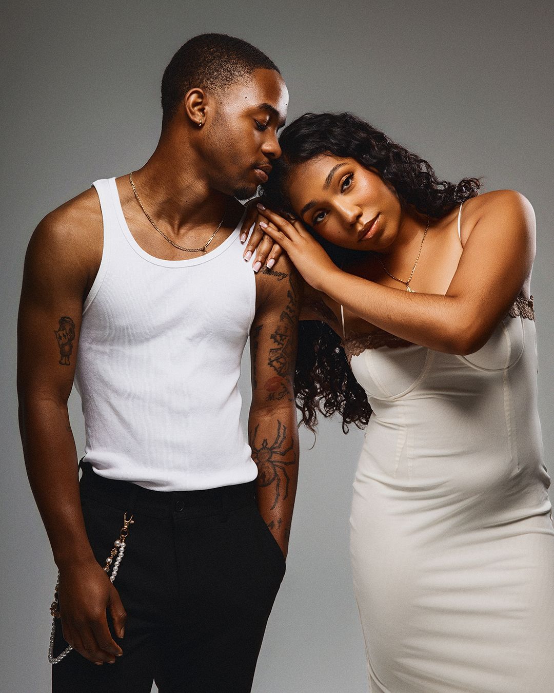 A stylish couple poses together, man in white tank top with tattoos, woman in white dress leaning on his shoulder.