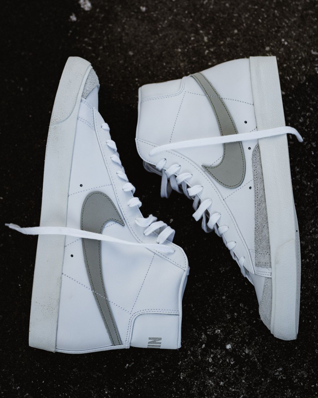 A pair of white Nike Blazer high-top sneakers with gray swoosh logos on a dark textured surface.