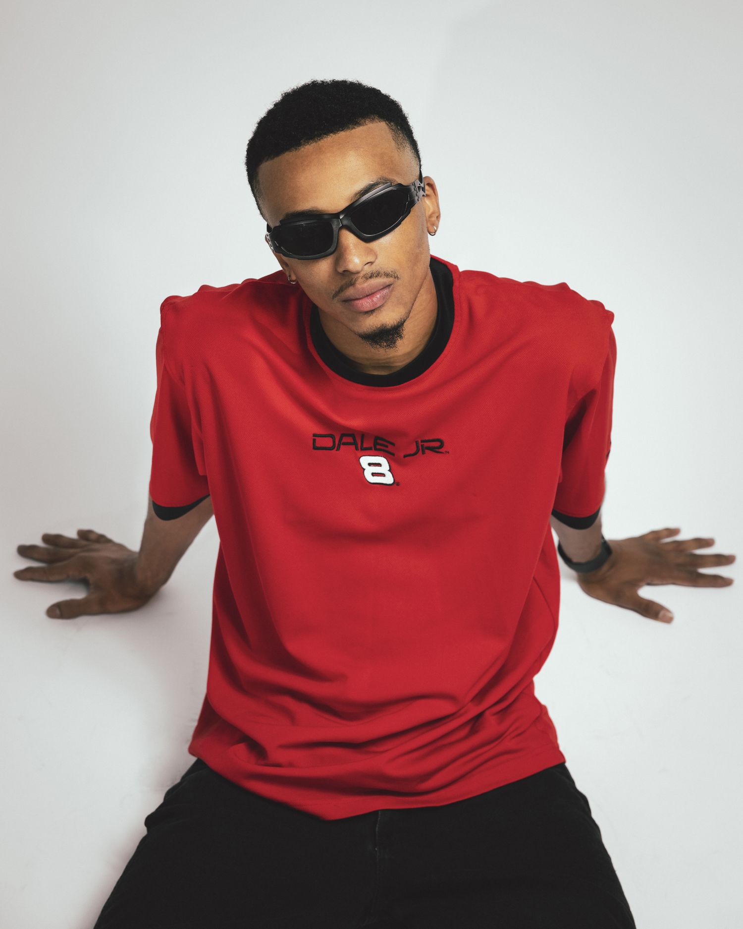 A person in a red t-shirt and black pants poses against a white background making expressive hand gestures.