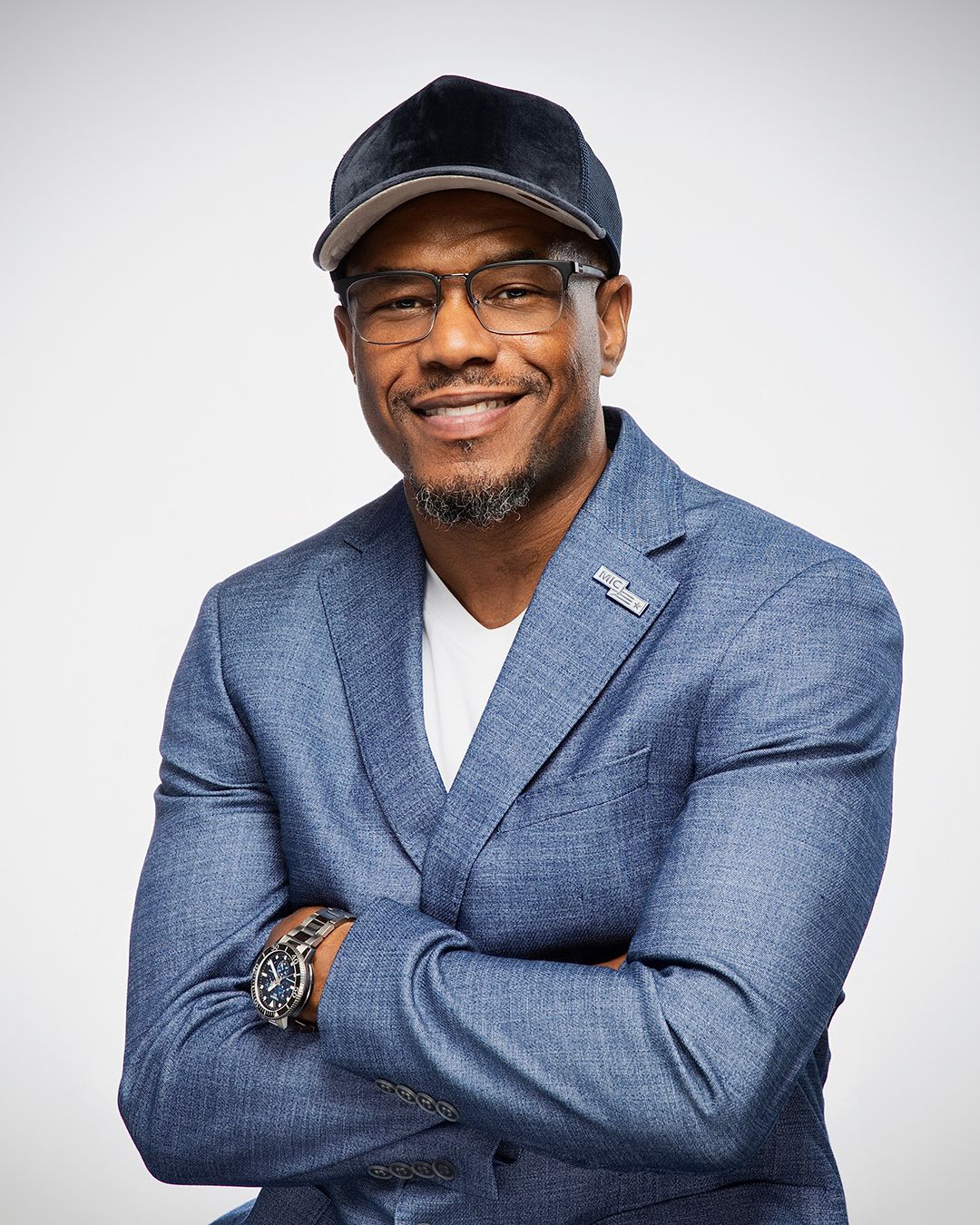 Confident man in blue blazer, cap, and glasses smiling with arms crossed against white background.