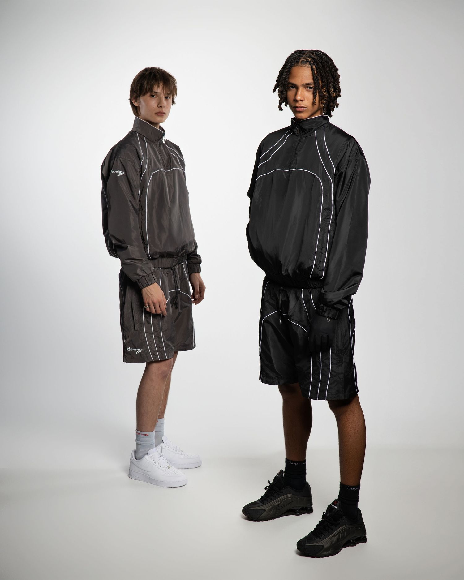 Models showcase dark oversized streetwear collection with white sneakers against minimalist studio backdrop.