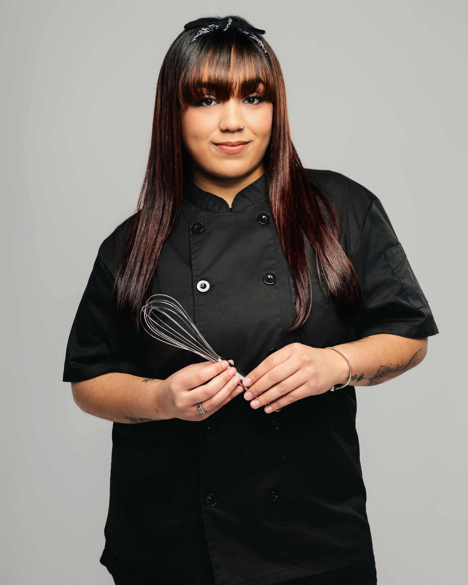 Professional chef in black uniform holding a whisk against gray studio background.