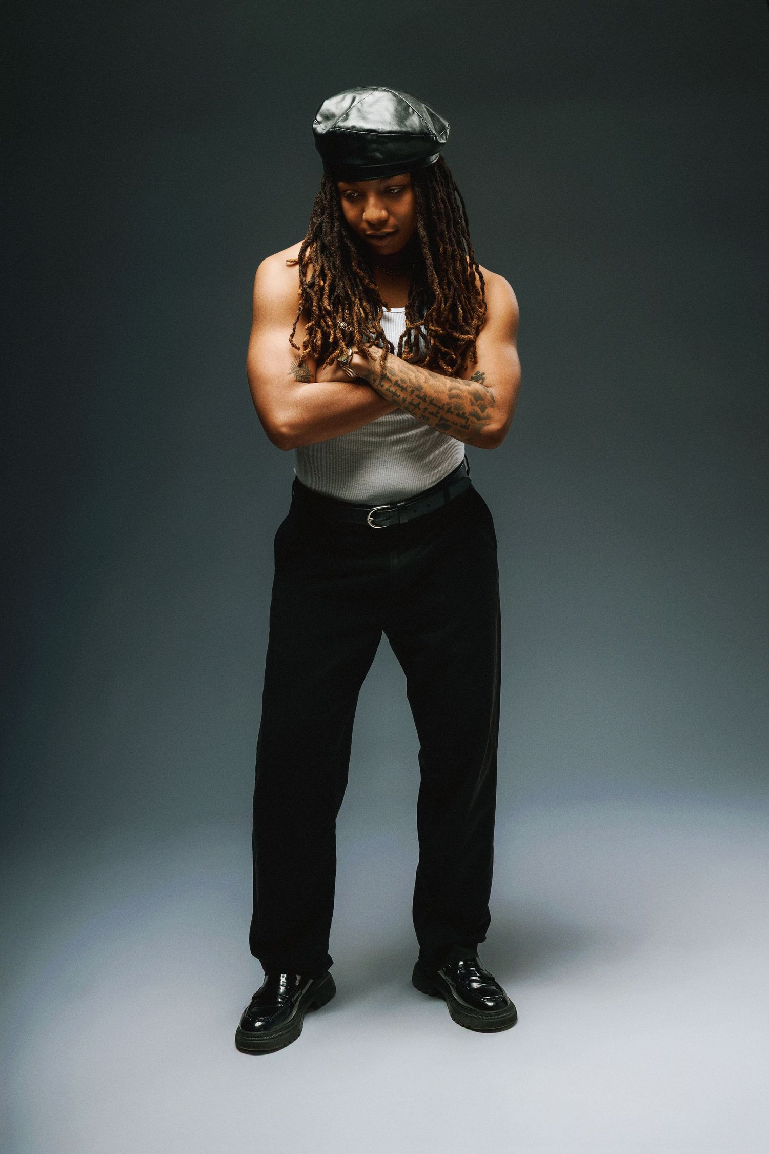Figure in tank top and black pants strikes confident pose with crossed arms wearing a newsboy cap against dark background.