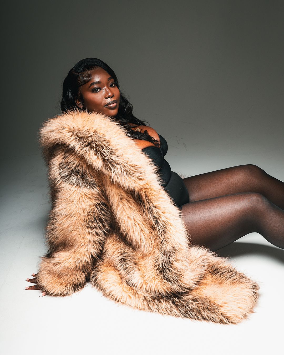 Woman in black bodysuit wearing a luxurious oversized tan fur coat, posing elegantly on a studio floor.