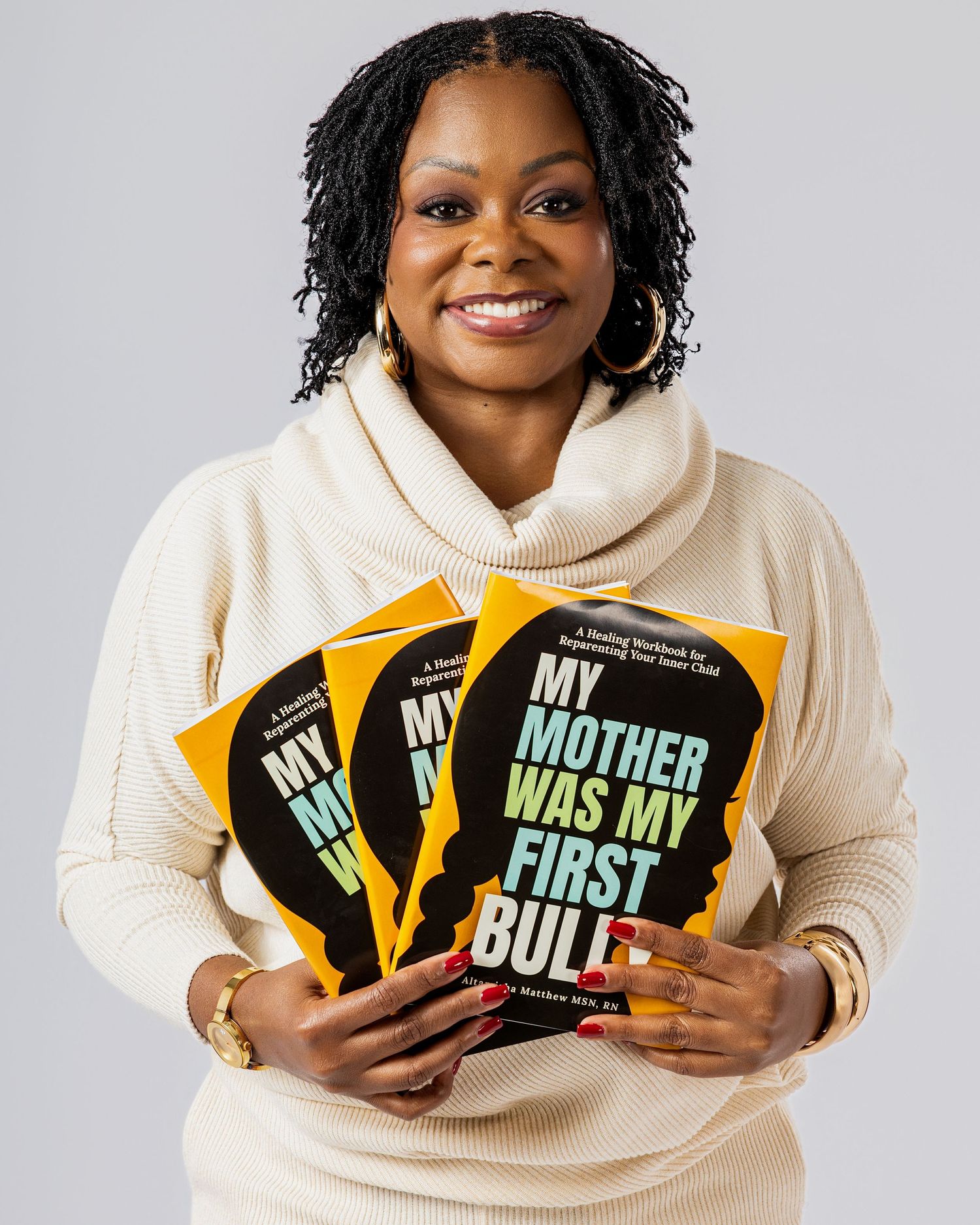 Person in cream hoodie holding multiple colorful books with text 'My Mother Was My First' visible on covers.