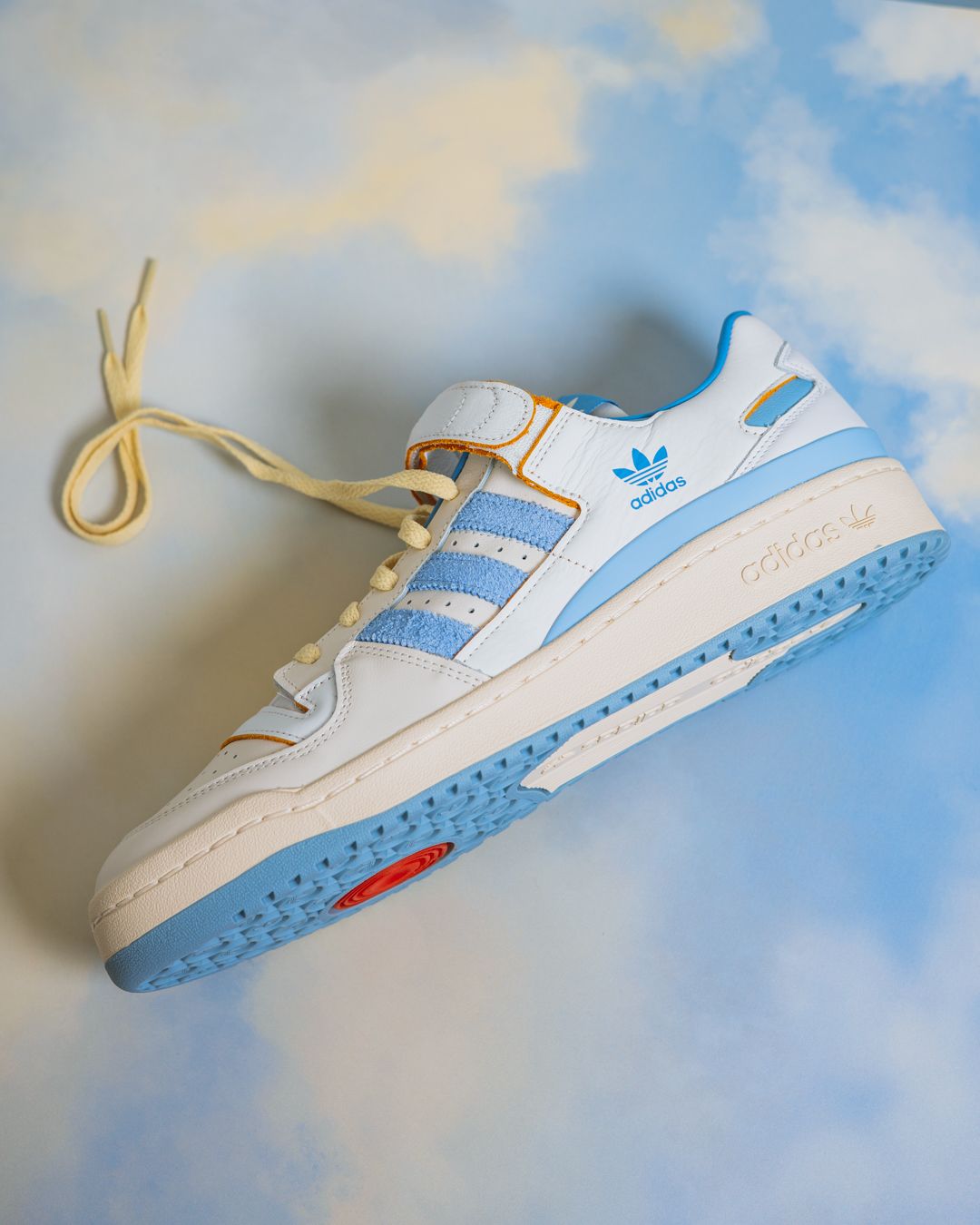 White and light blue Adidas sneaker with cream laces photographed against a cloudy sky background.