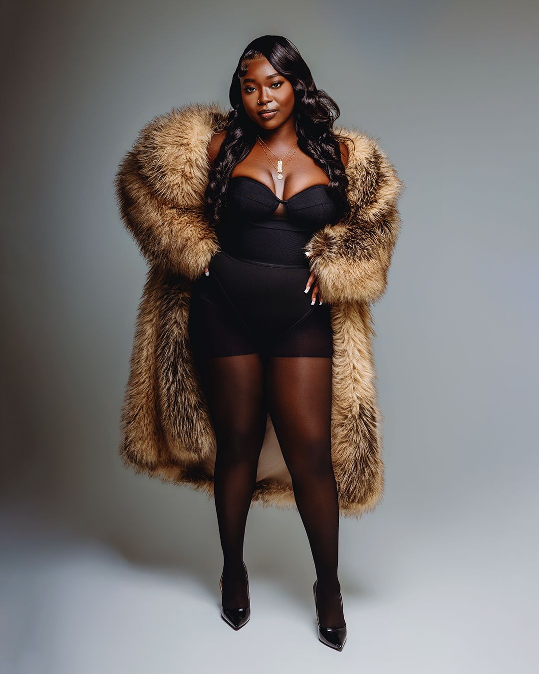 Woman in black bodysuit, sheer tights, heels, and oversized brown fur coat poses confidently in studio.