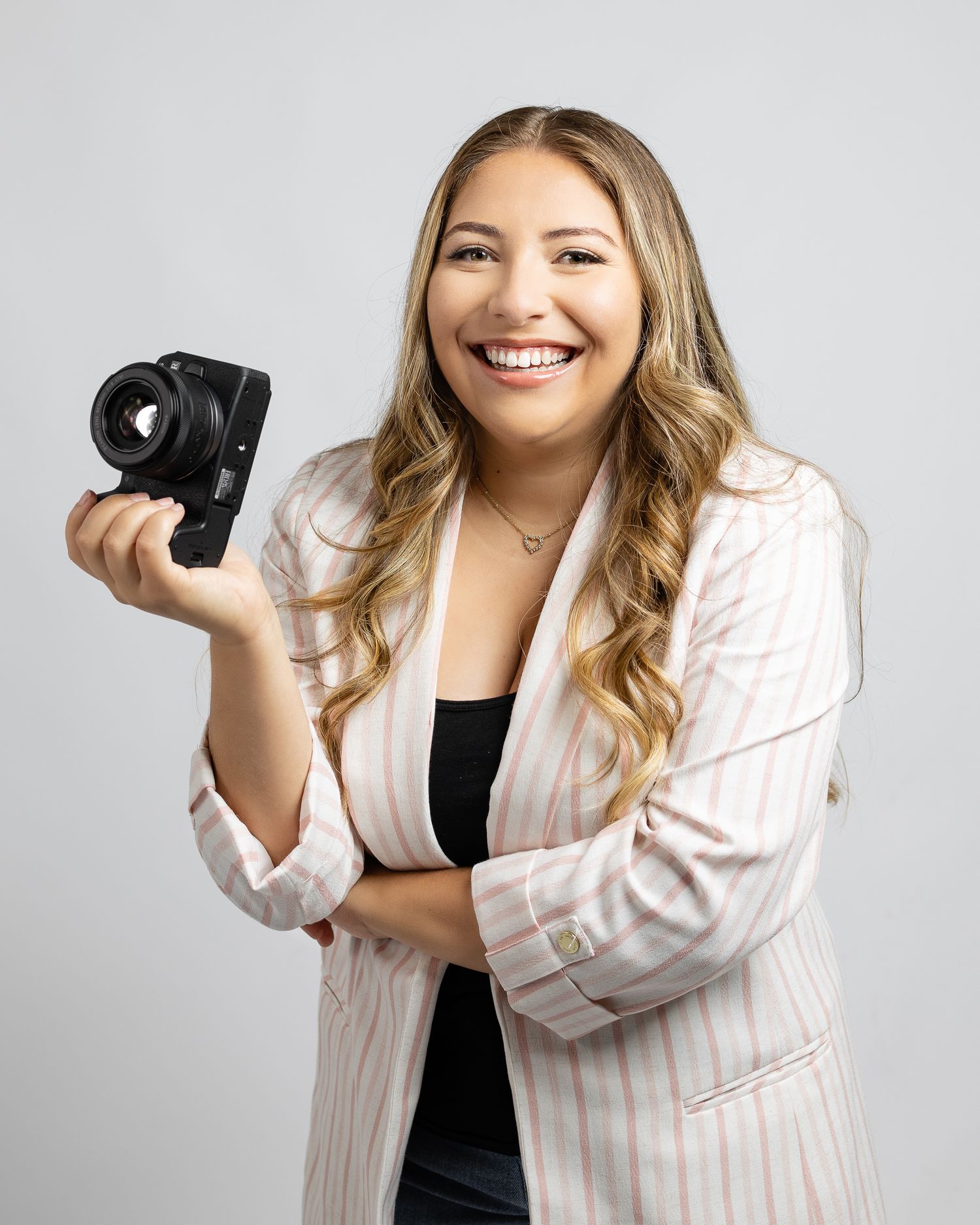 Professional photographer in pink striped blazer and black top poses with camera against white background.