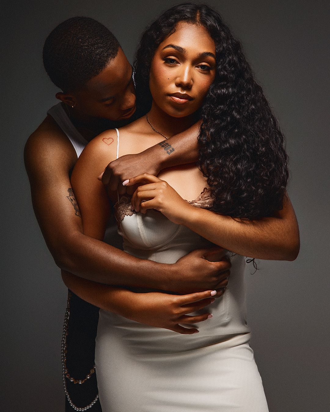 A couple in an intimate embrace, woman in white dress with long curly hair, man holding her from behind, studio portrait.