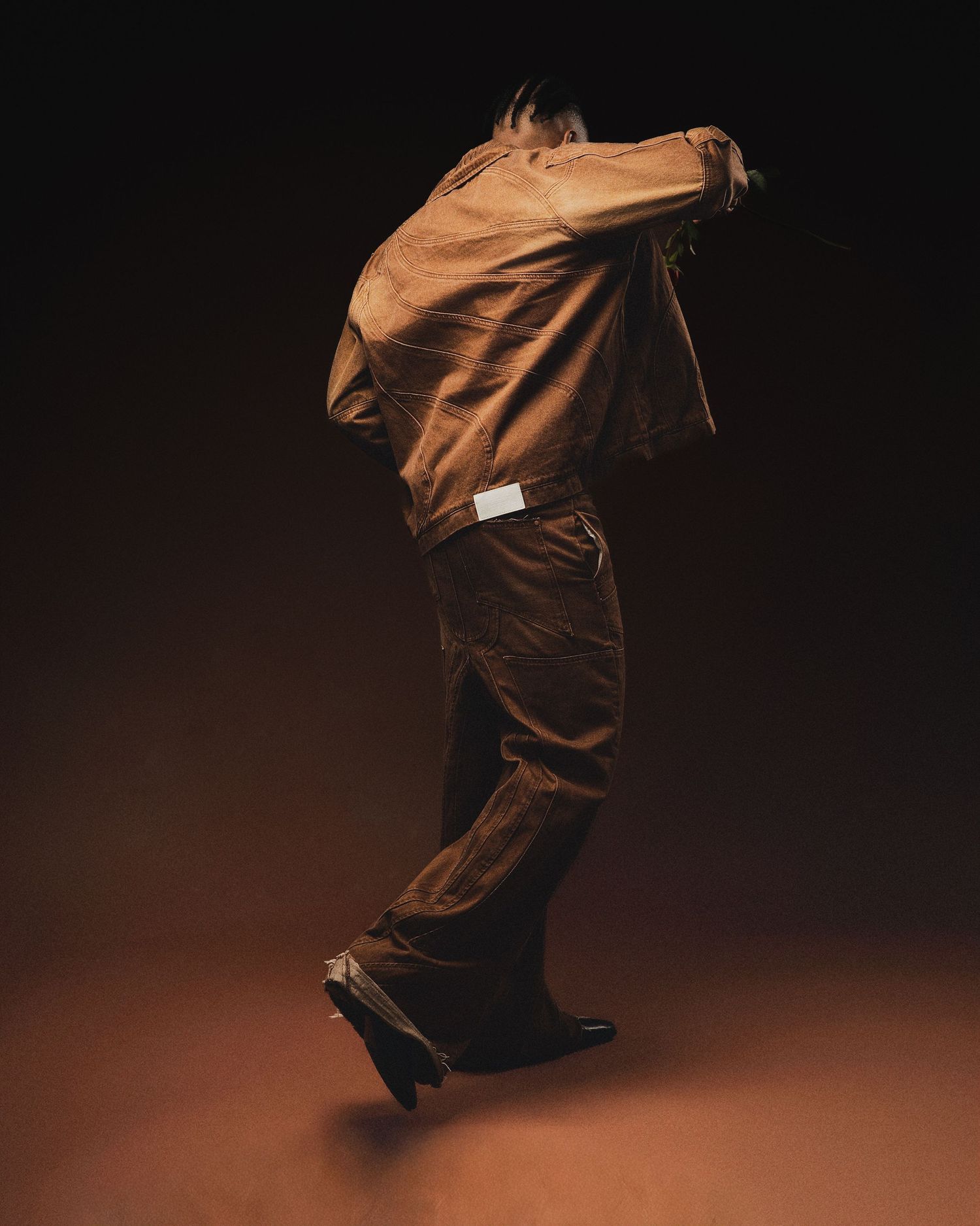 Dynamic dance pose showing figure in brown outfit against dark studio backdrop.
