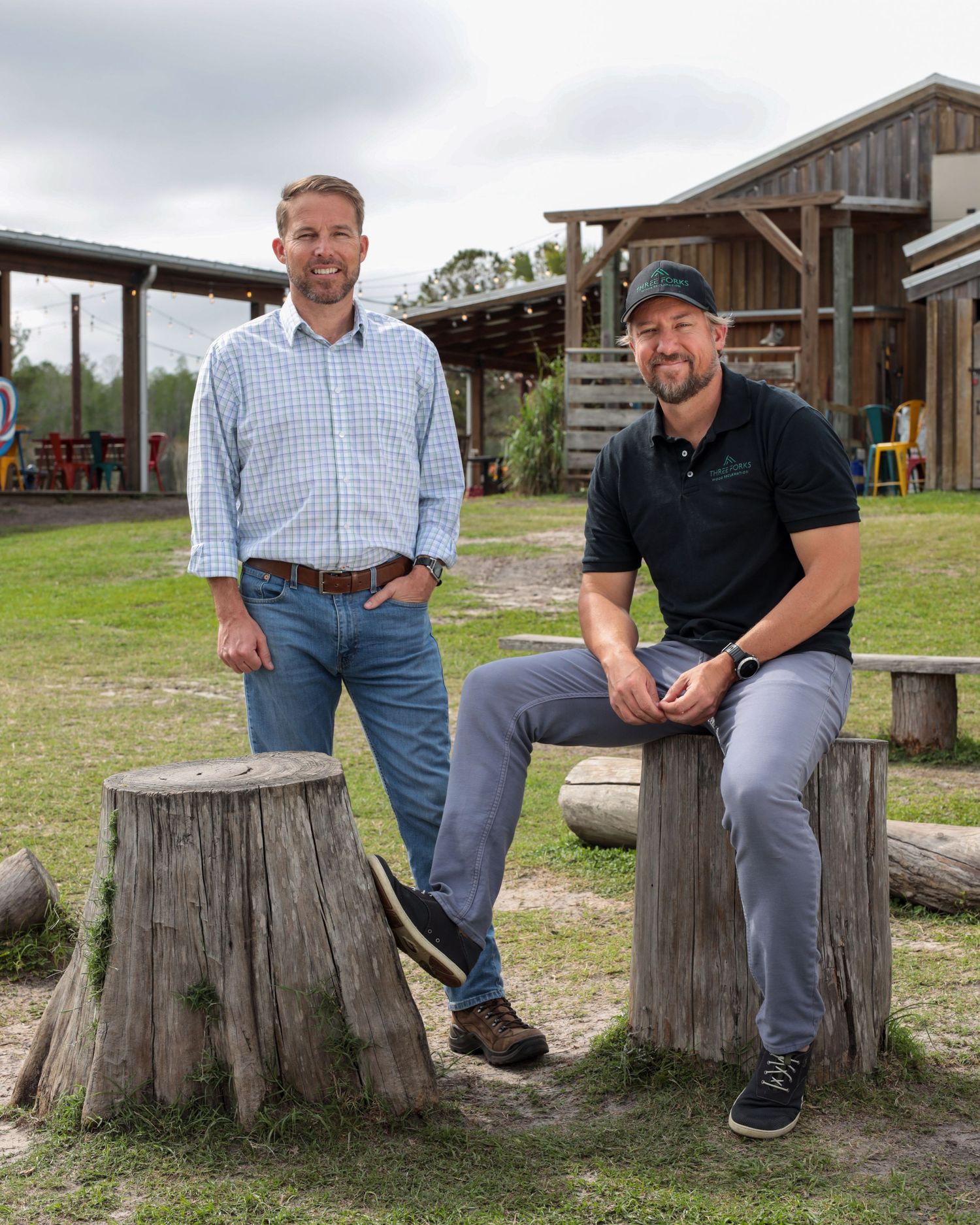Two men in casual attire sit on tree stumps in front of a rustic barn structure on a grassy field.