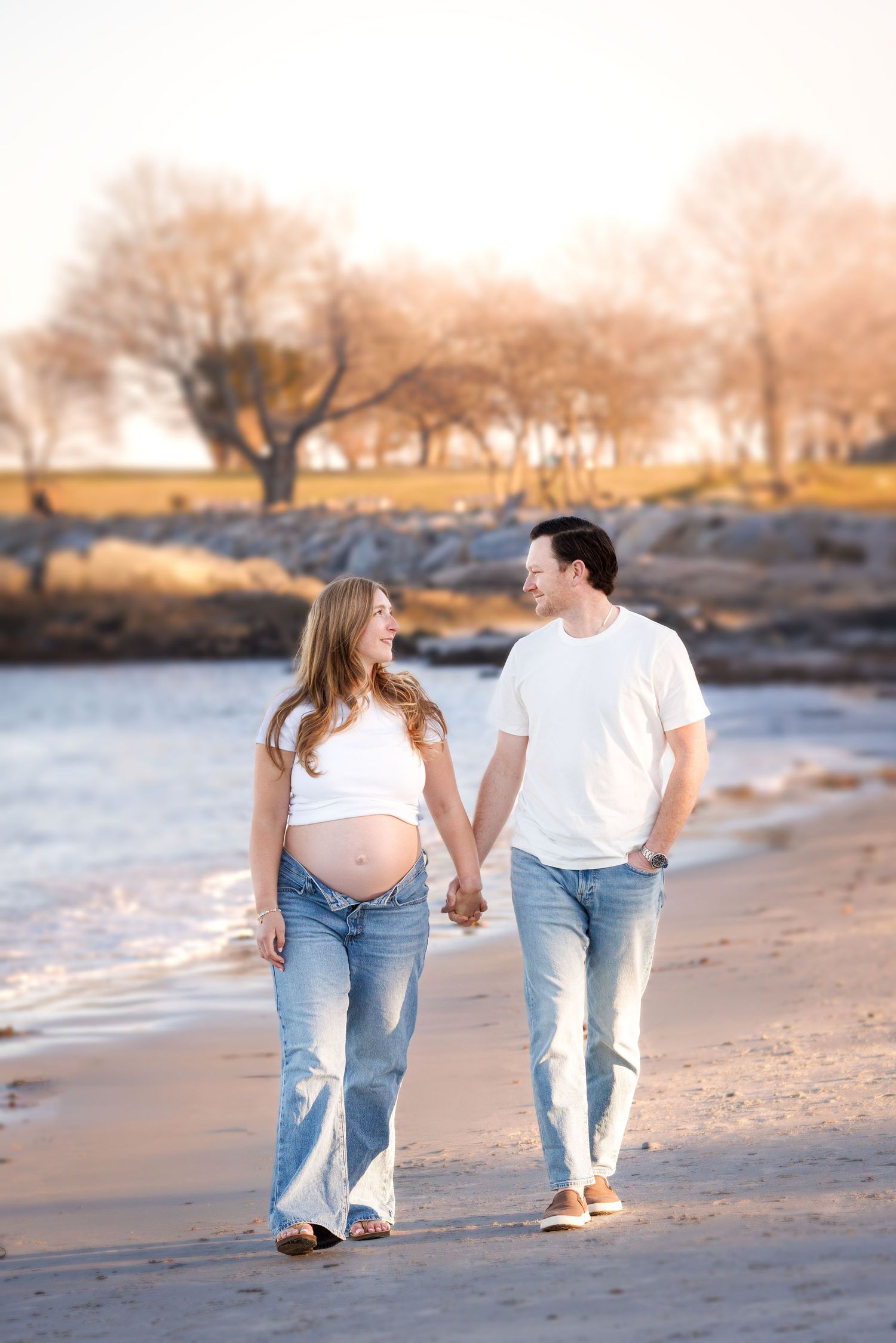 Pregnant couple holding hands walking on a sandy beach at sunset, wearing white tops and jeans.