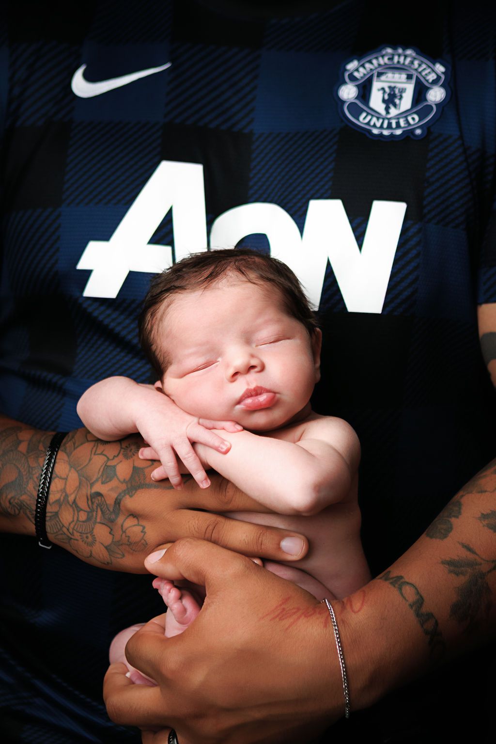 Sleeping newborn baby cradled in tattooed hands against a Manchester United AON jersey.