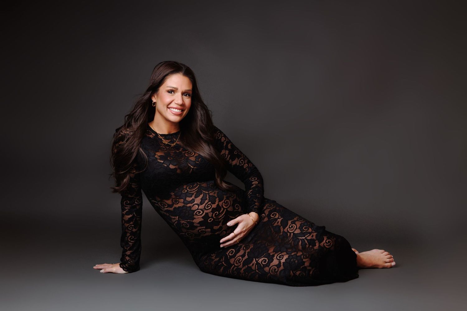 Pregnant woman in black lace maternity gown posing gracefully on floor against gray studio background.