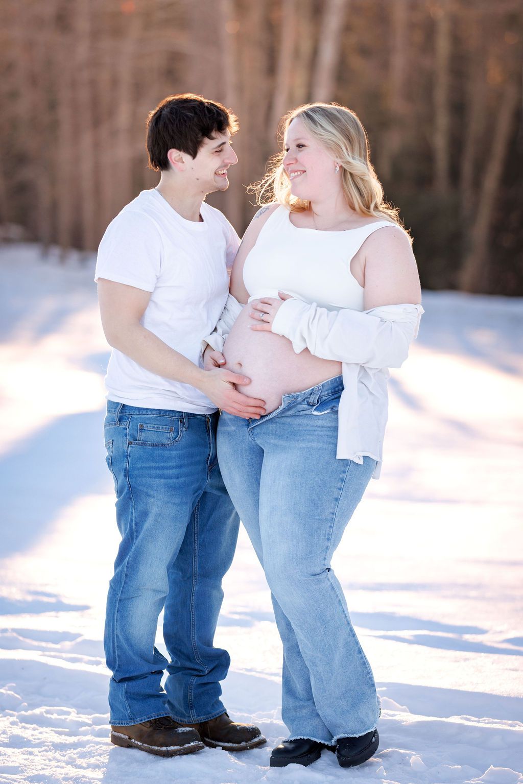 Couple sharing a tender moment in snowy winter setting, woman showing pregnant belly, both wearing white tops and jeans.