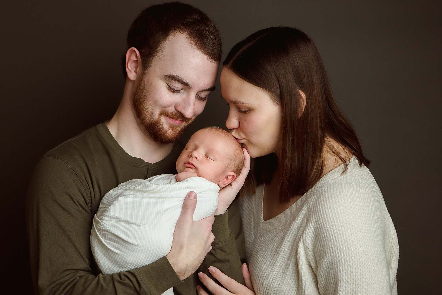 Parents lovingly embrace their sleeping newborn baby wrapped in white swaddle against dark background.