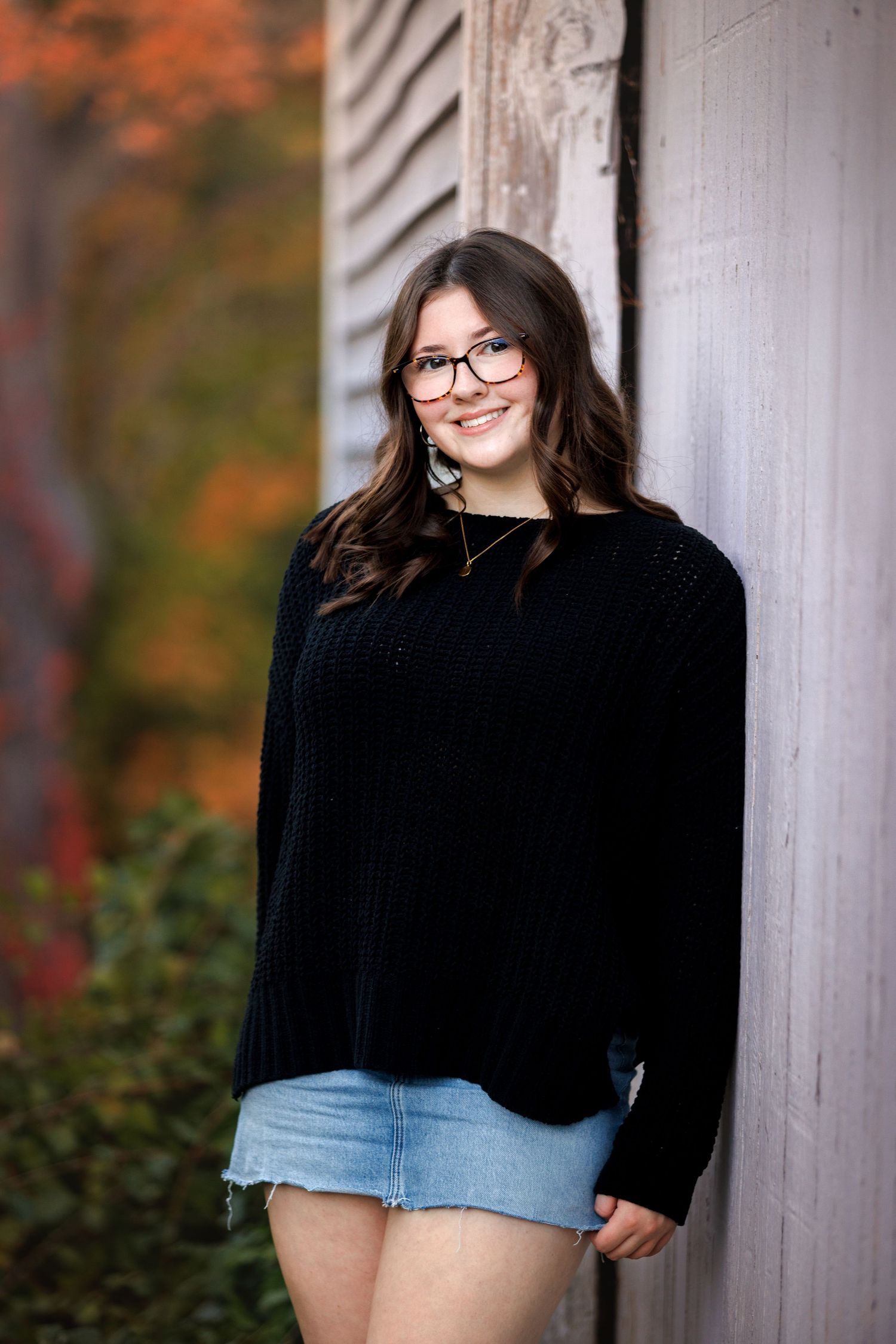 Senior girl wearing a black sweater and denim shorts poses against a white pillar with autumn foliage in the background.