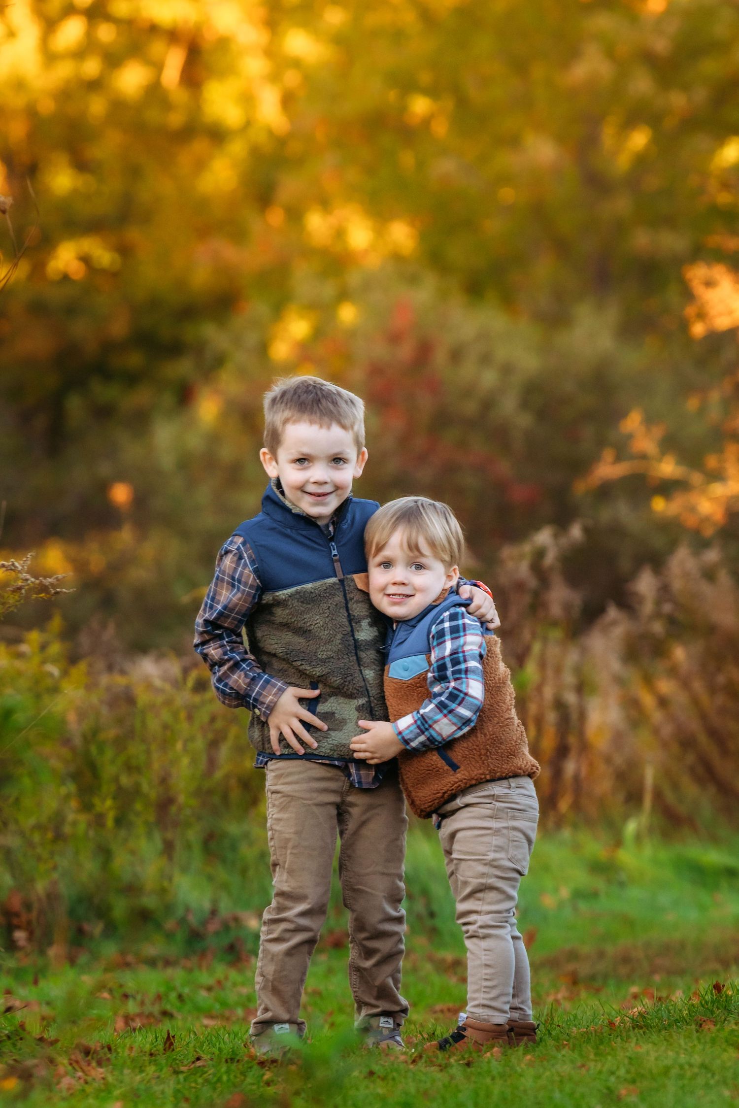Two children in plaid shirts and brown pants play together in a grassy autumn field at sunset.